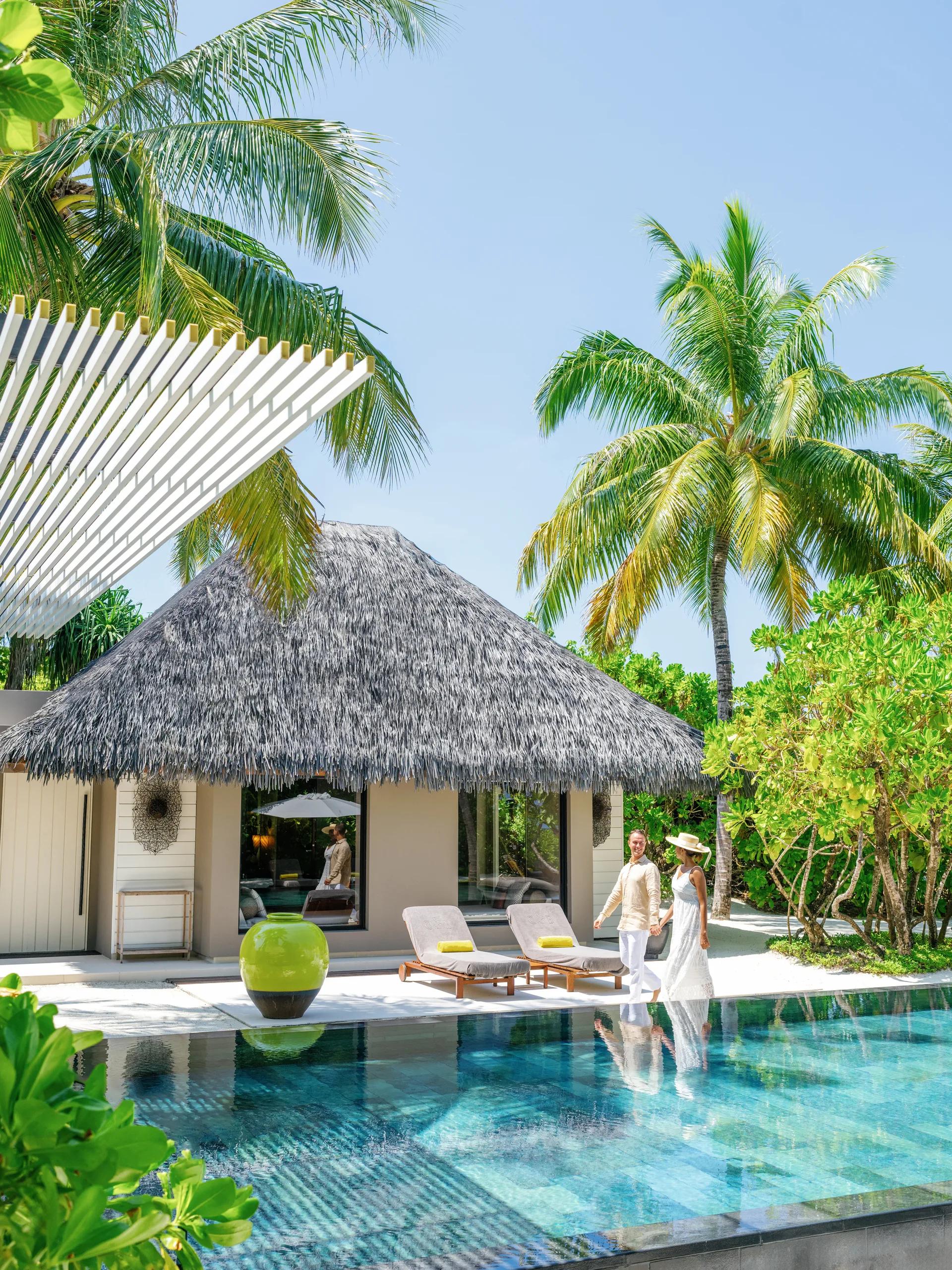 A couple walking beside an Island Villa's private pool, thatched roof and garden visible behind them