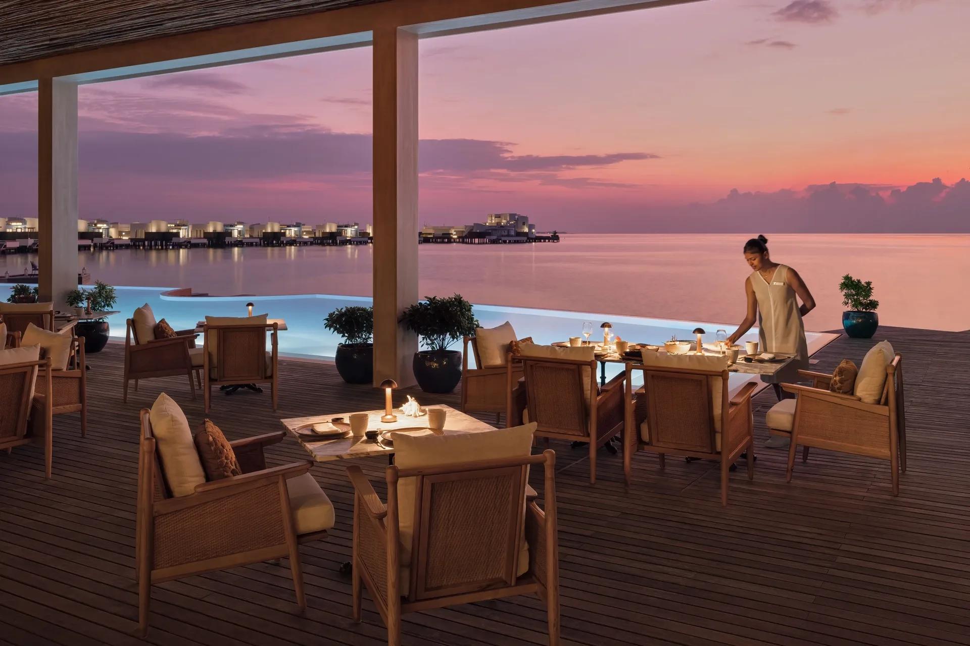 A dining terrace at dusk beside an infinity pool, overwater villas visible across the water — Alaaya Bay has no dedicated photograph in the source material; this stands in as the closest available shoreline dining scene