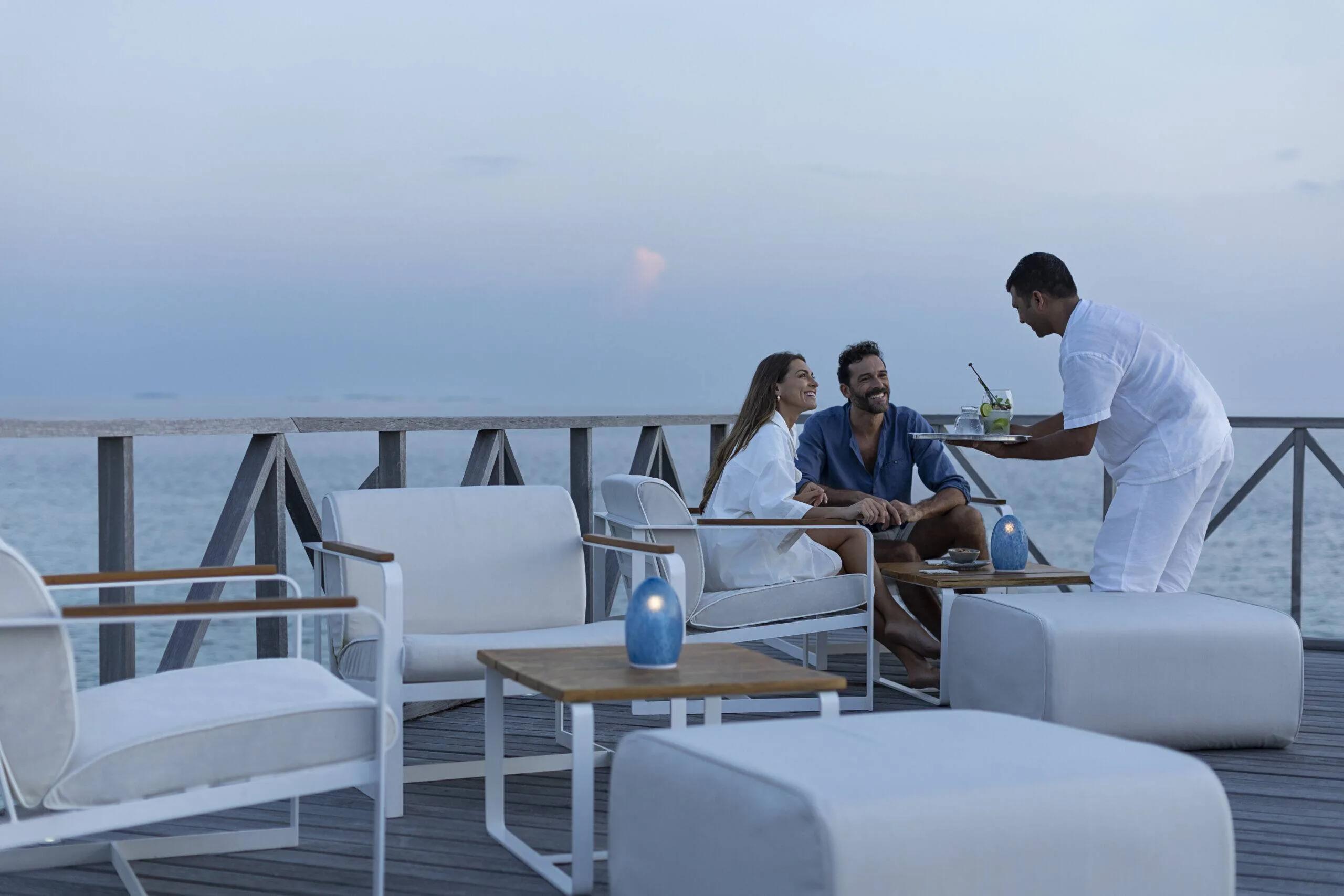 A waiter serving drinks to a couple relaxing on Raw Sunset Deck's lounge seating at dusk