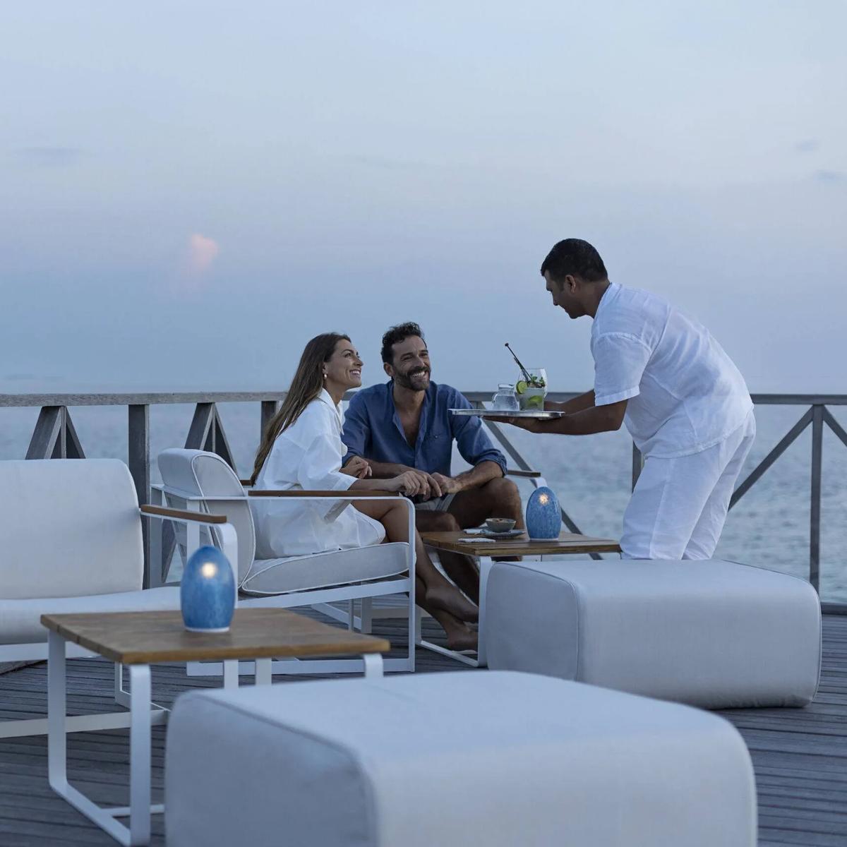 A waiter serving drinks to a couple relaxing on Raw Sunset Deck's lounge seating at dusk