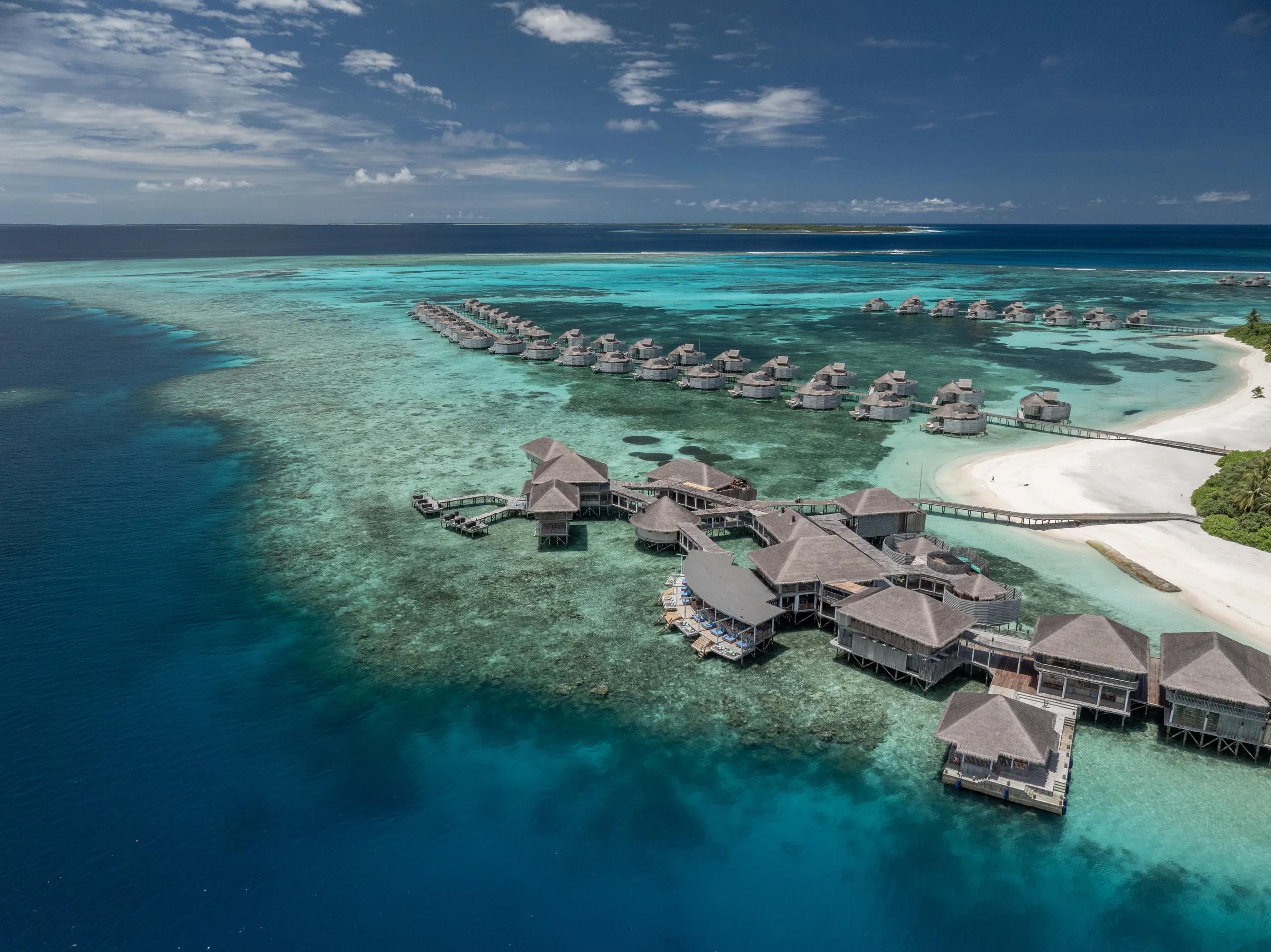 Aerial view of Six Senses Laamu's overwater villas strung along the reef edge, with the main jetty building and a white-sand beach on Olhuveli Island beyond