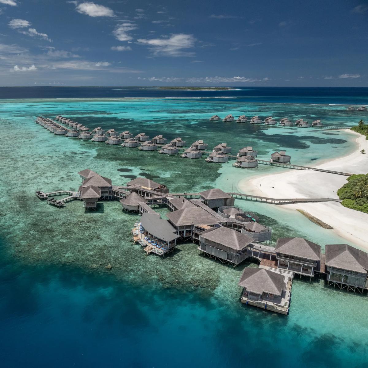 Aerial view of Six Senses Laamu's overwater villas strung along the reef edge, with the main jetty building and a white-sand beach on Olhuveli Island beyond