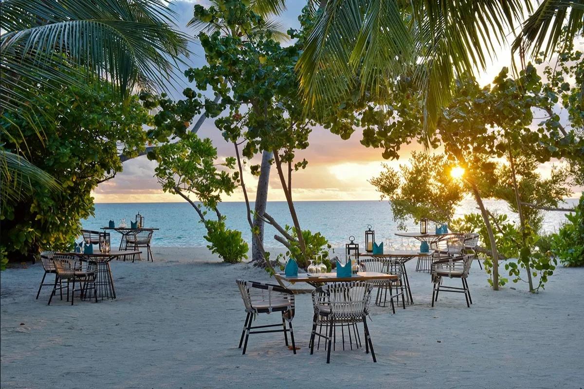 A private table set on the beach at sunset, glassware and lanterns laid among the trees