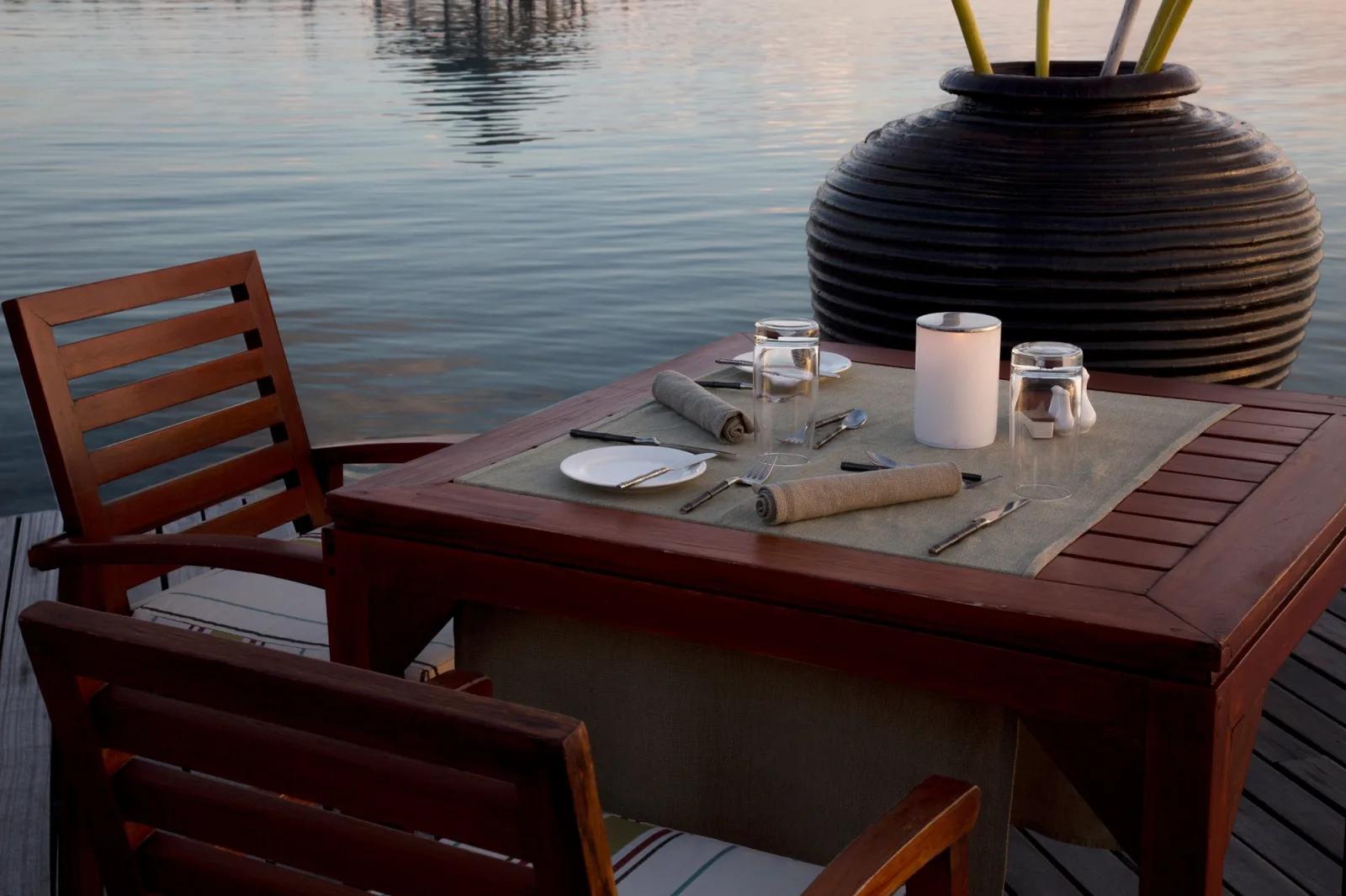 A table for two set at the water's edge at dusk, large ceramic urn beside the deck