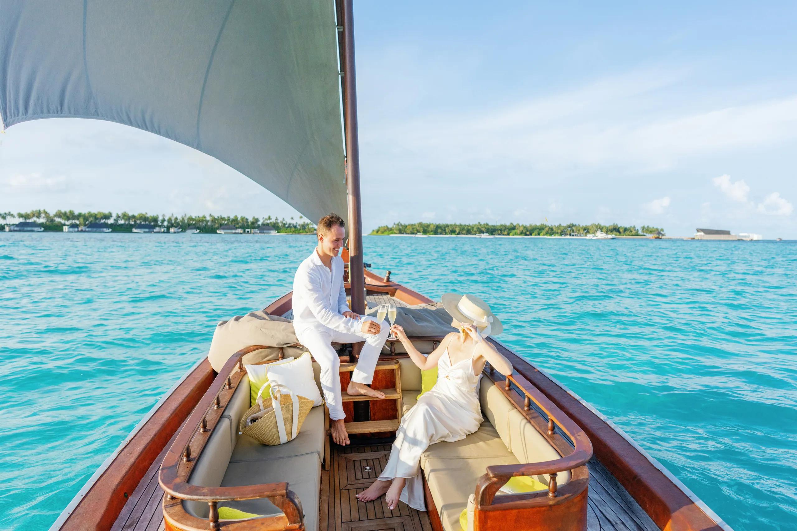 A couple toasting champagne aboard a traditional wooden dhoni under sail, neighboring islands visible across the lagoon