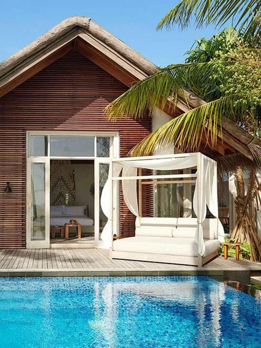 A larger beach villa exterior with private pool, canopied daybed, and thatched roofline, set among palms