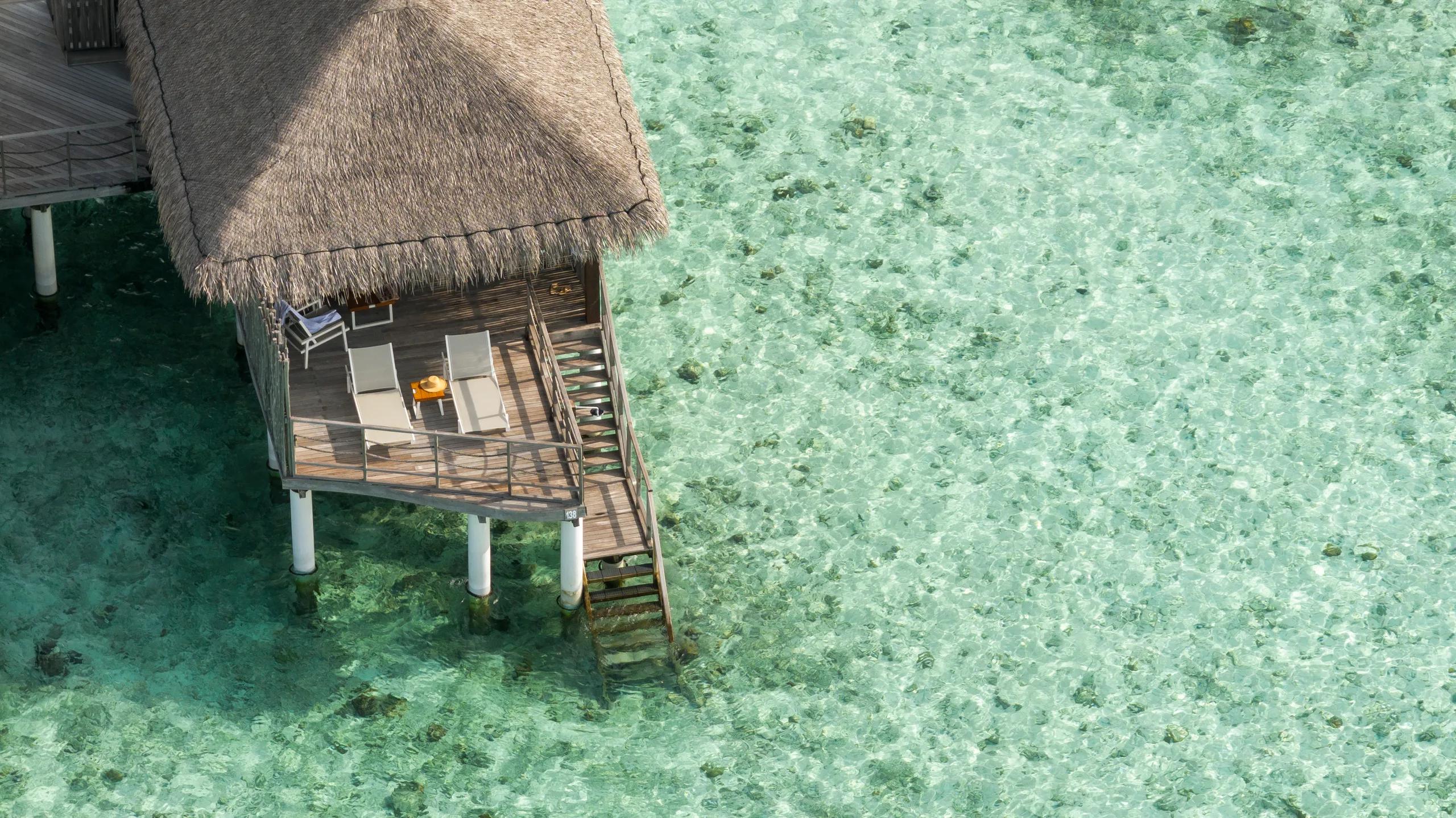 Aerial view of a single Water Villa's thatched roof and sun-deck standing on stilts over clear reef water