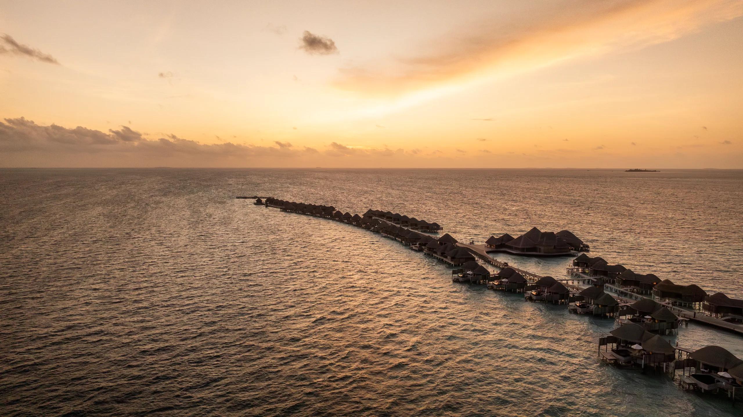 Aerial view at sunset of Constance Halaveli's overwater villas stretching along a single jetty into open water, with a small island visible on the horizon
