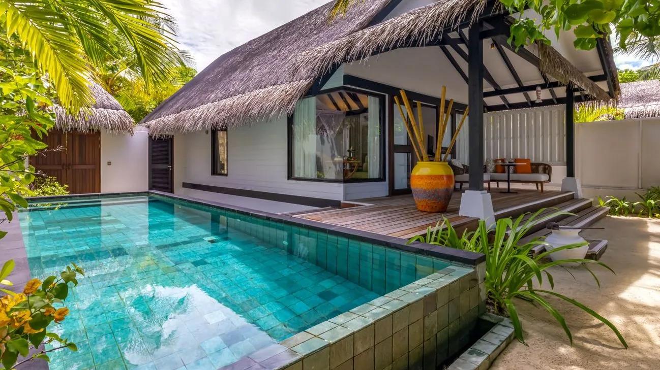 A beach villa's private pool bordered by dark tile, the thatched-roof villa and deck behind it