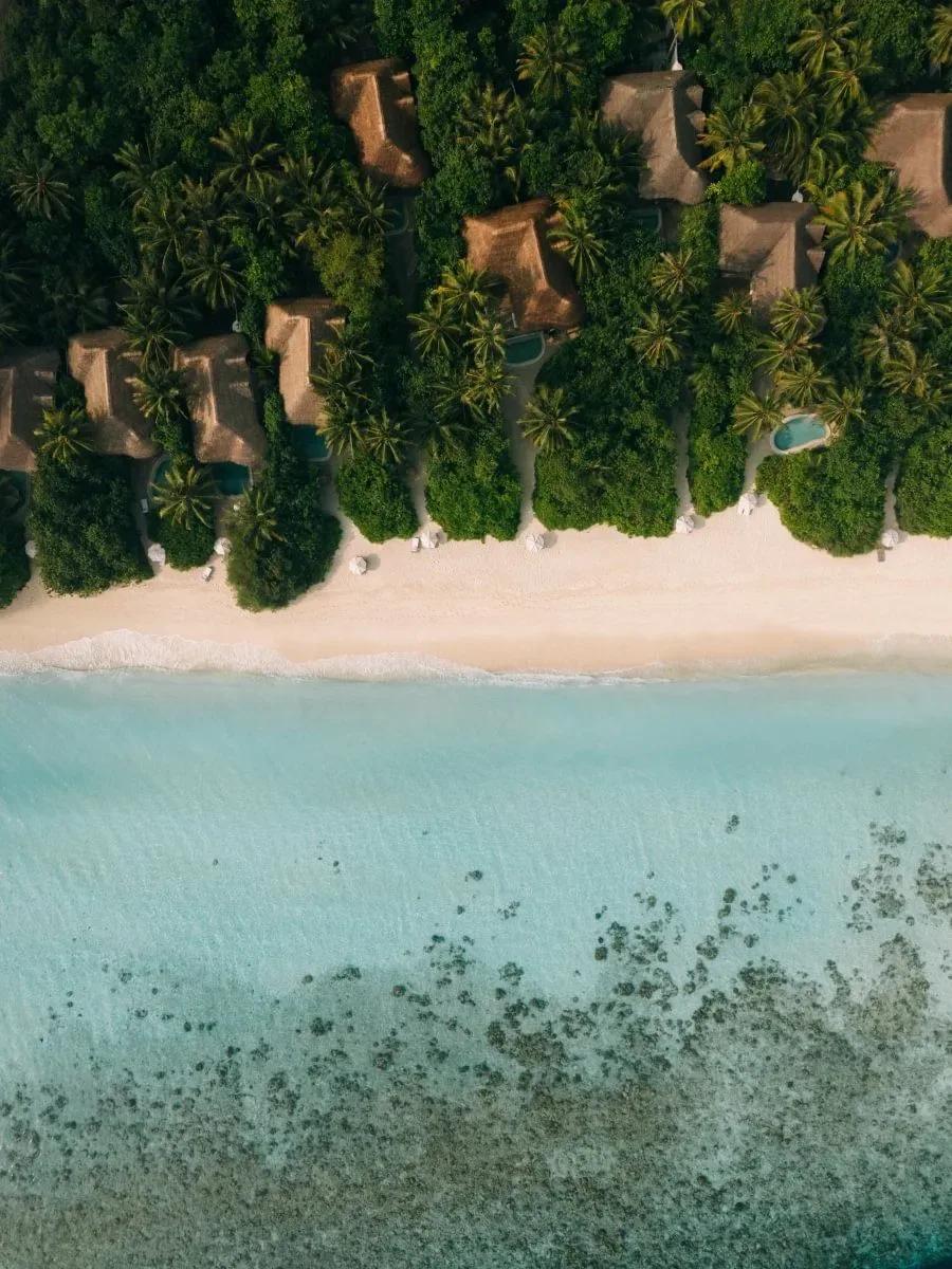 A directly-overhead aerial view of Kunfunadhoo Island's shoreline — thatched villa roofs and private pools set among dense jungle, above a white-sand beach where the reef drops into open water
