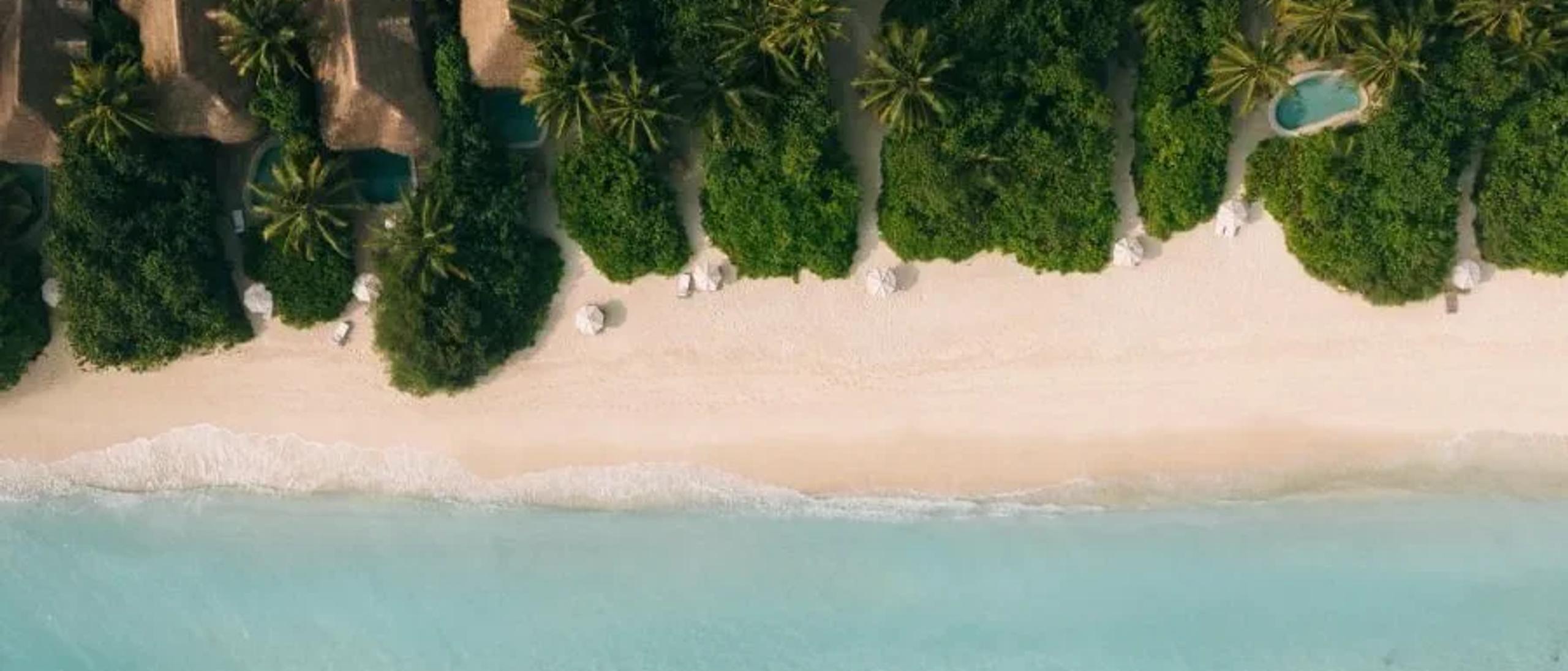 A directly-overhead aerial view of Kunfunadhoo Island's shoreline — thatched villa roofs and private pools set among dense jungle, above a white-sand beach where the reef drops into open water