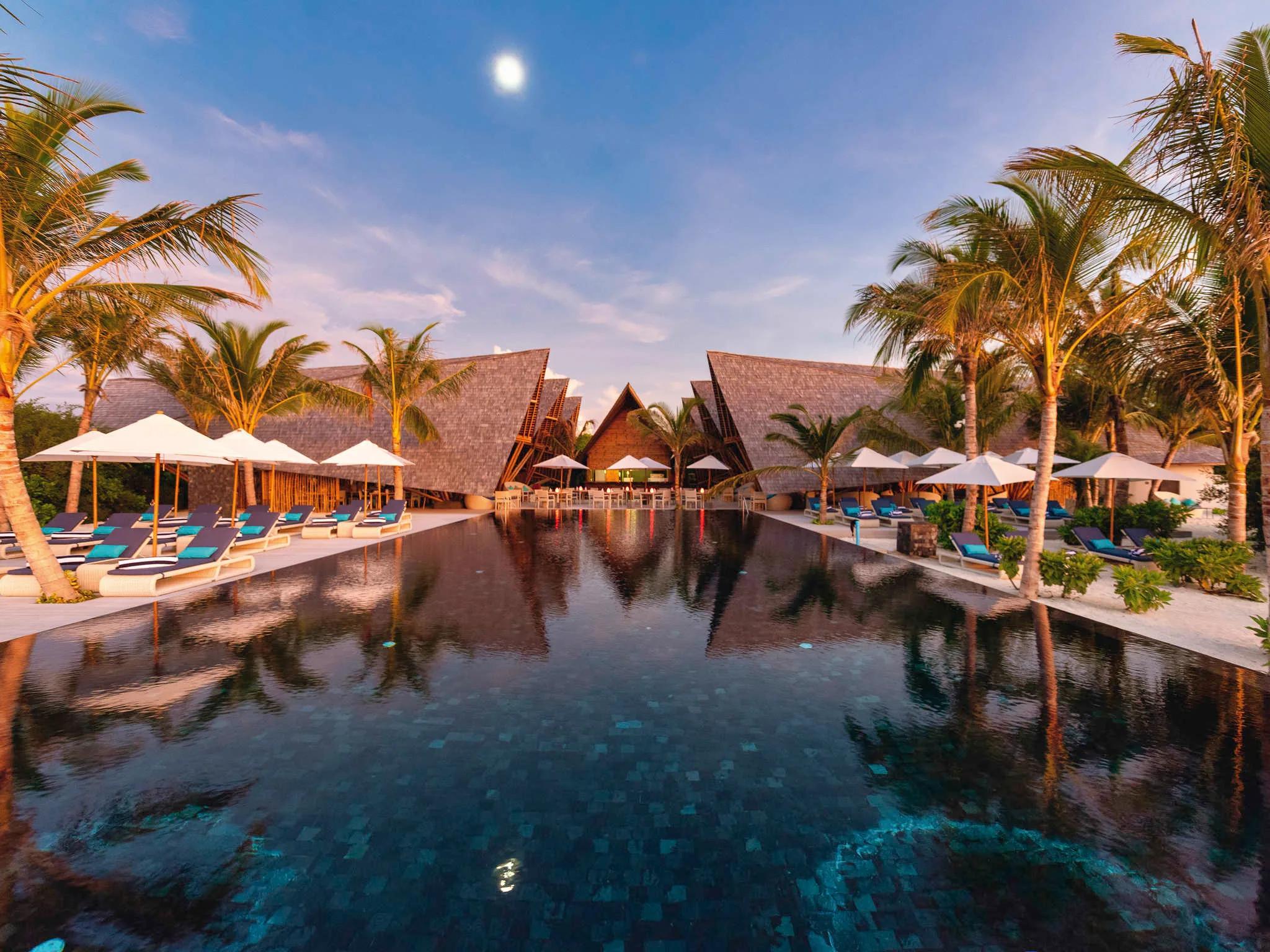 Latitude 5.5's poolside dining terrace at dusk, angular thatched roofs lit above rows of loungers either side of the reflecting pool