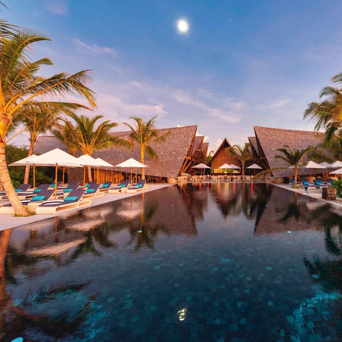 Latitude 5.5's poolside dining terrace at dusk, angular thatched roofs lit above rows of loungers either side of the reflecting pool