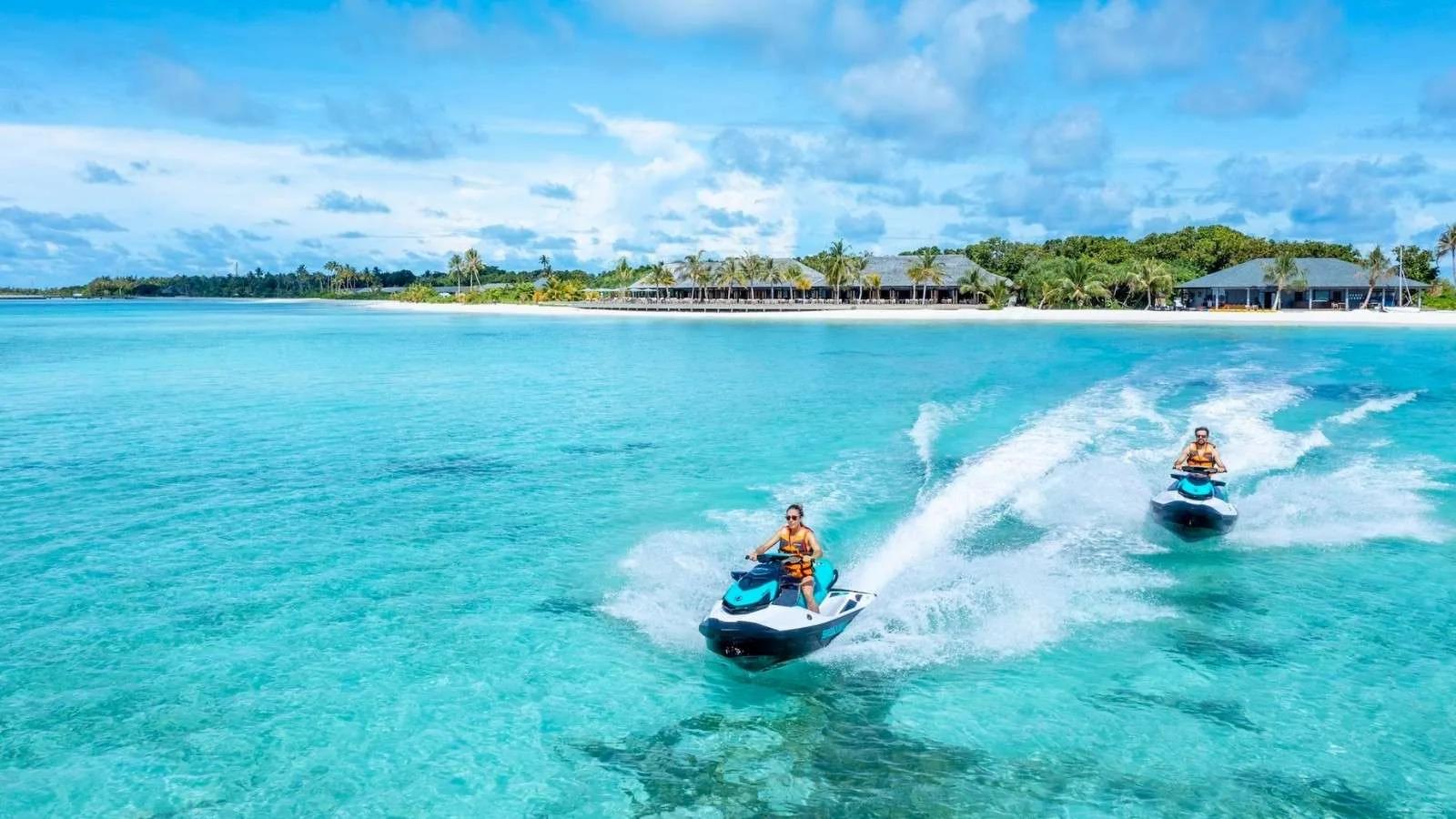 Two guests riding jet skis across the lagoon at Jawakara, the resort's beachfront villas visible behind them