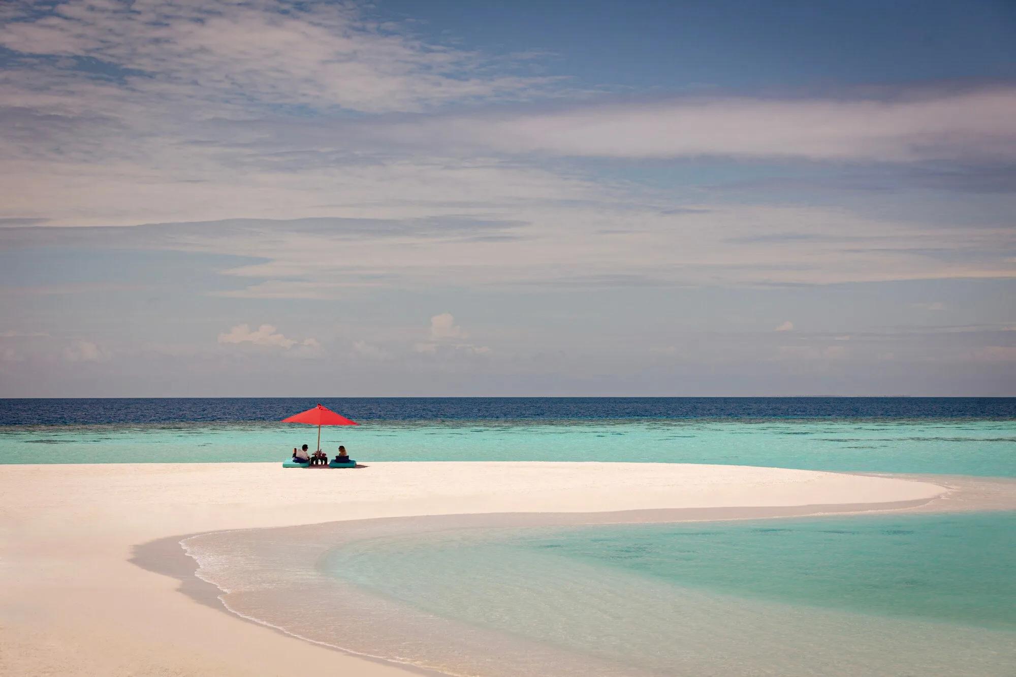 A couple seated under a single red umbrella on an empty white sandbank, reef and open ocean stretching out on either side
