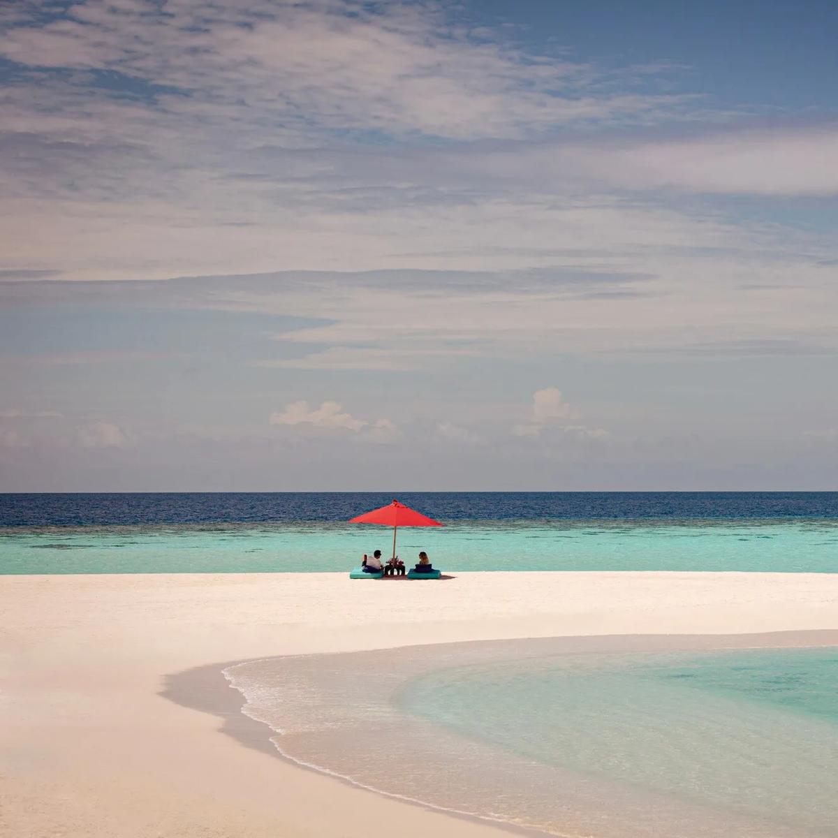 A couple seated under a single red umbrella on an empty white sandbank, reef and open ocean stretching out on either side