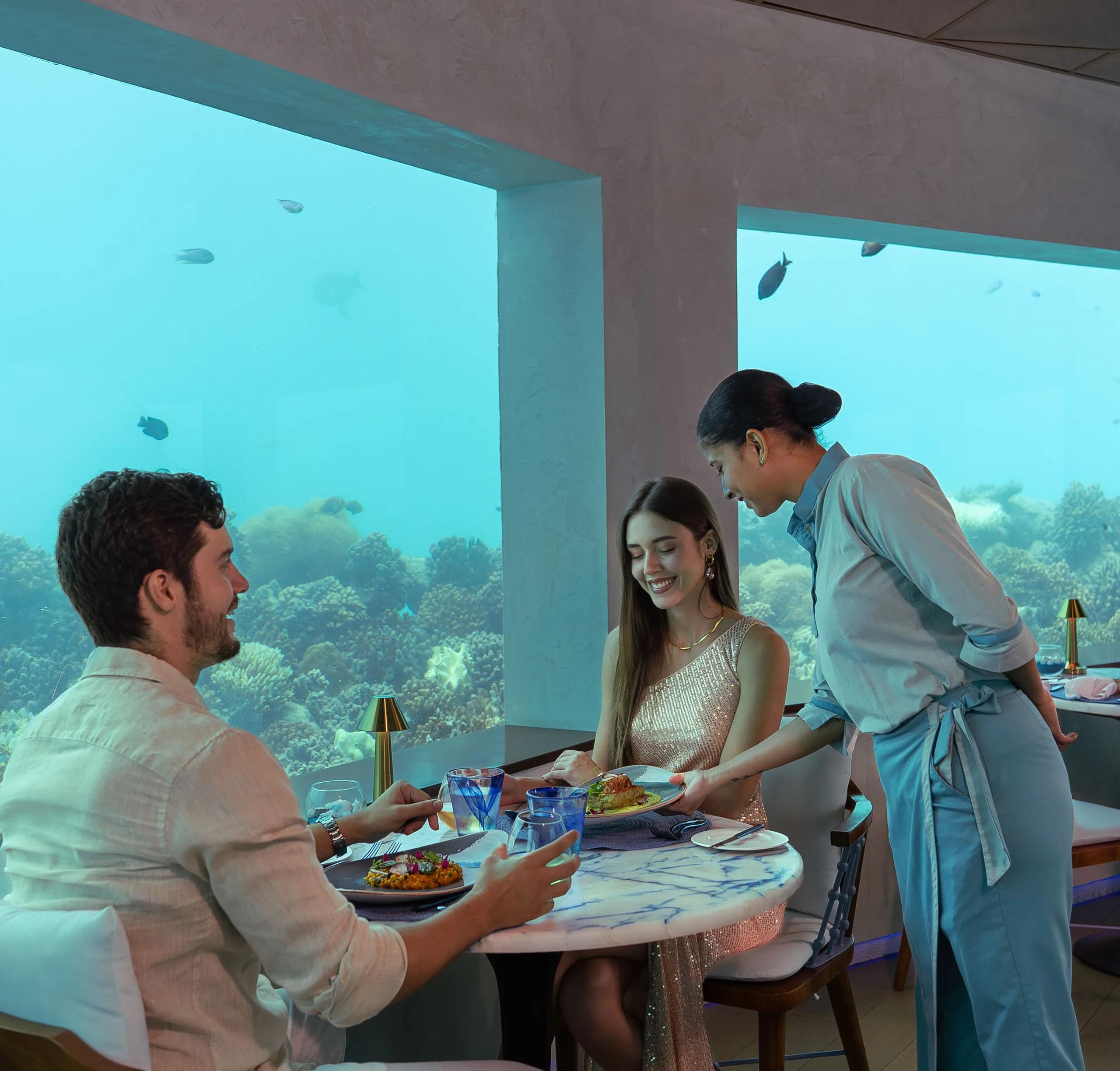 A couple dining at Only BLU, OBLU SELECT Lobigili's under-ocean restaurant, reef visible through floor-to-ceiling windows on both sides
