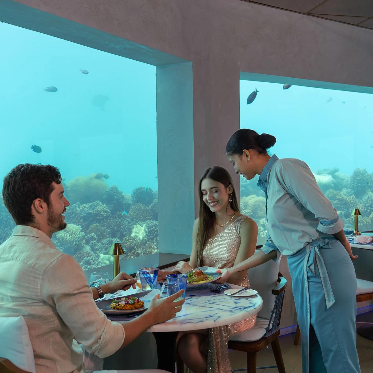 A couple served dinner at Only BLU, OBLU SELECT Lobigili's under-ocean restaurant, coral reef visible through floor-to-ceiling windows
