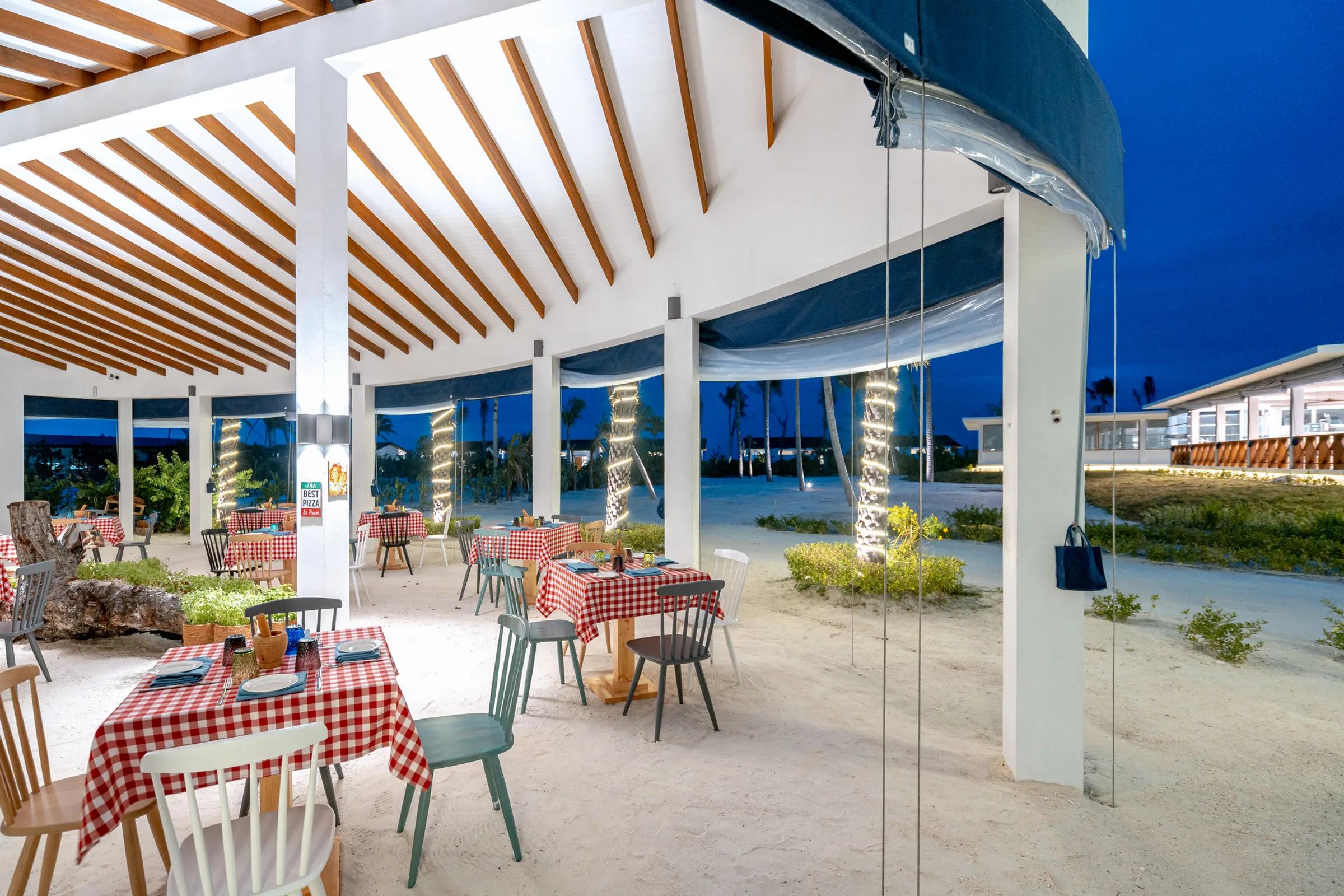 Il Basilico's beachside dining room at dusk, red-checked tablecloths set beneath a timber pergola strung with lights