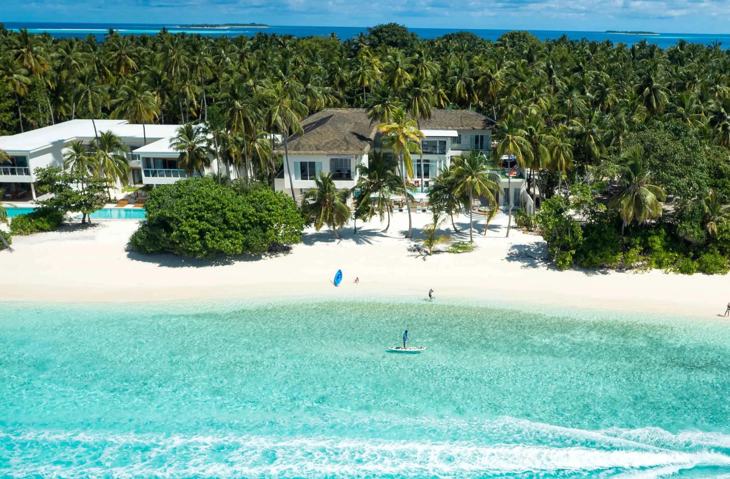 A beach residence seen from above through the palms at Amilla, with a paddleboarder and kayak in the shallow lagoon