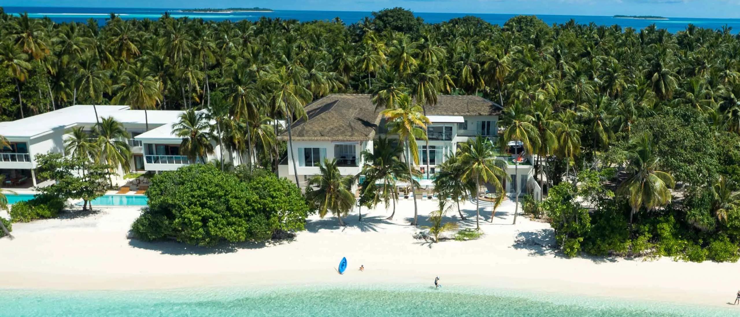 A beach residence seen from above through the palms at Amilla, with a paddleboarder and kayak in the shallow lagoon