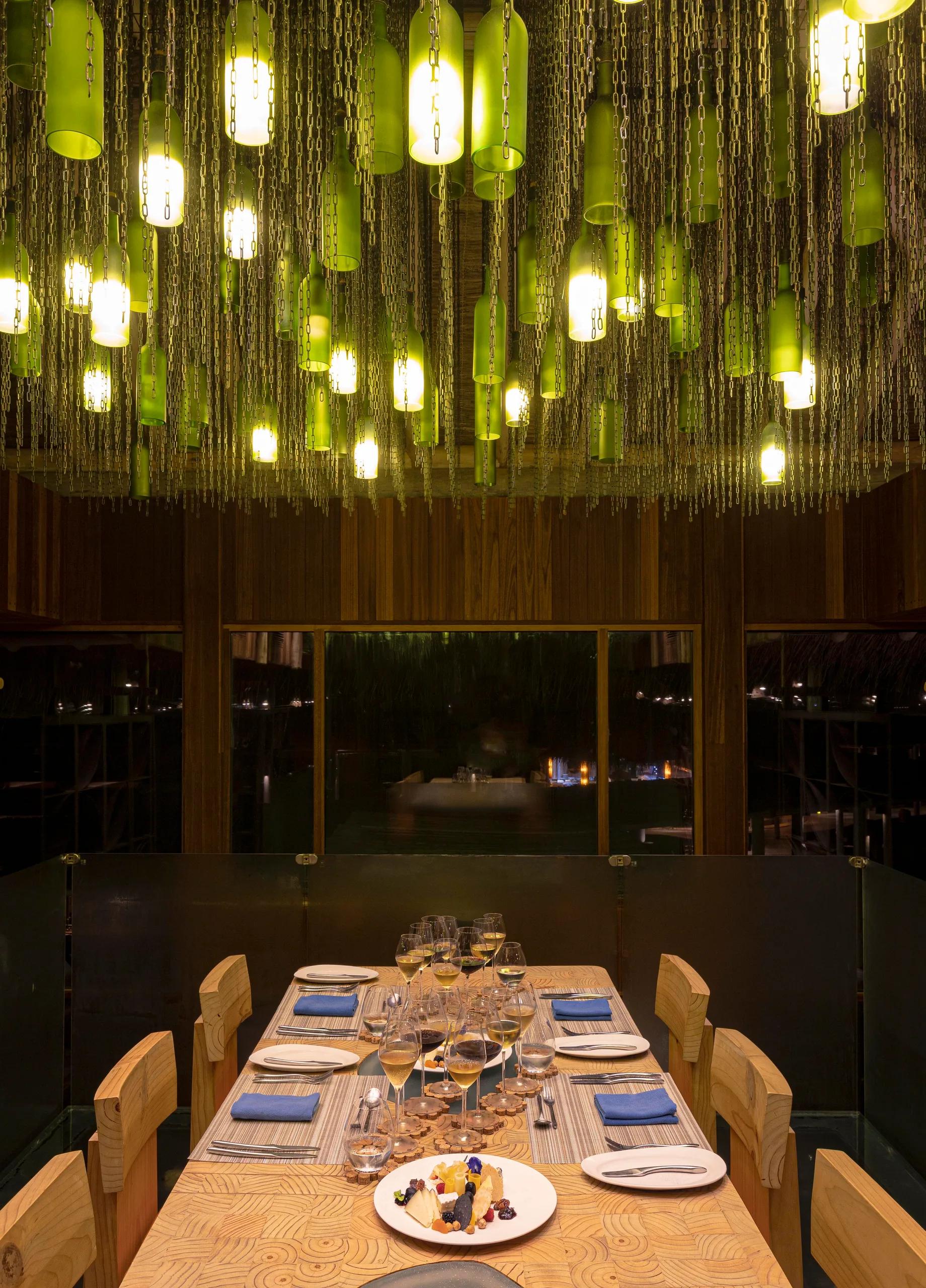 Green wine bottles hang on chains as chandelier lighting above a private dining table set with glasses