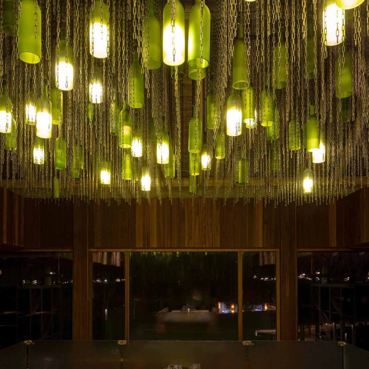 Green wine bottles hang on chains as chandelier lighting above a private dining table set with glasses