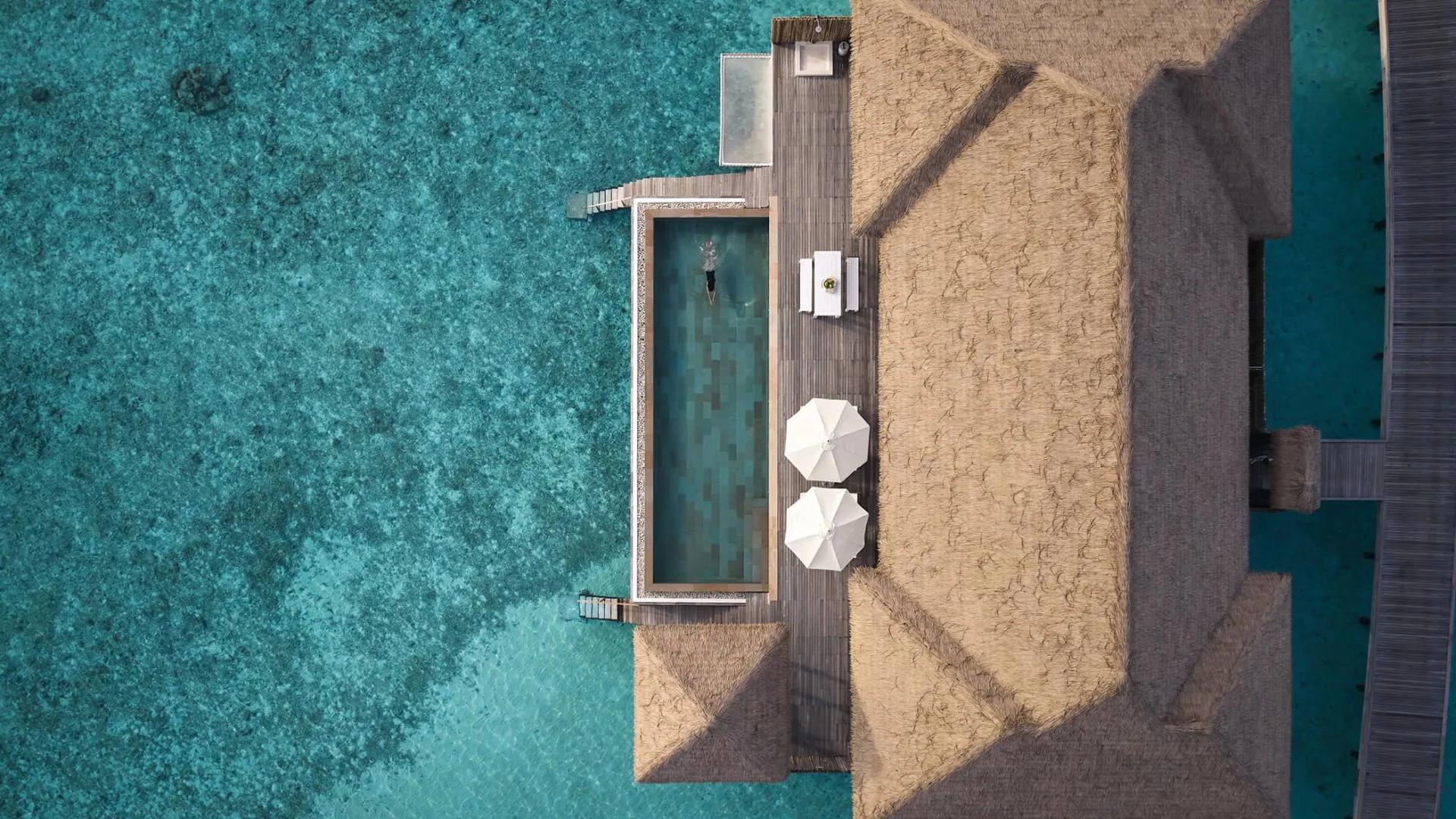 Aerial view directly above an overwater villa's thatched roof and private pool, with a swimmer in the pool and open lagoon on either side
