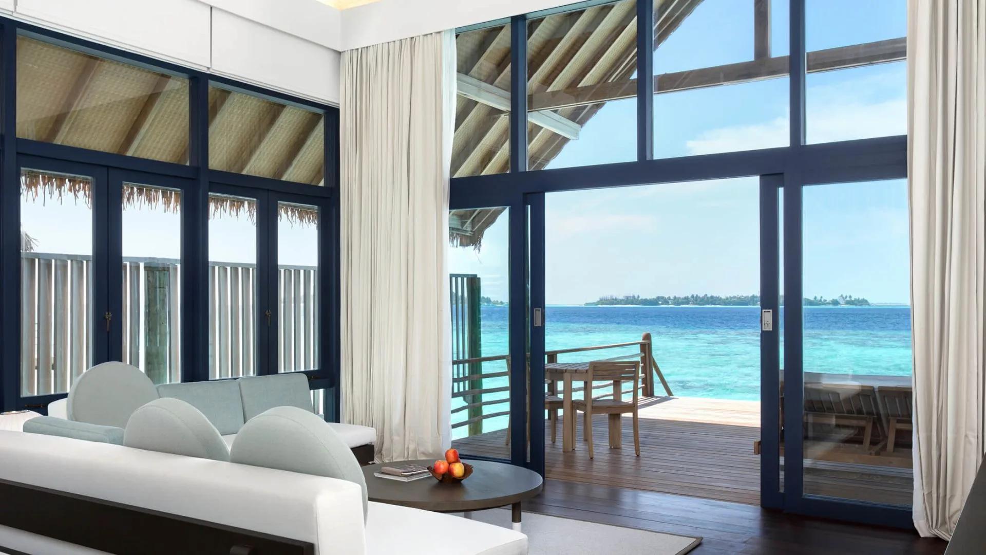 The living area of a Loft Water Villa, glass doors open onto a private overwater deck and the lagoon beyond