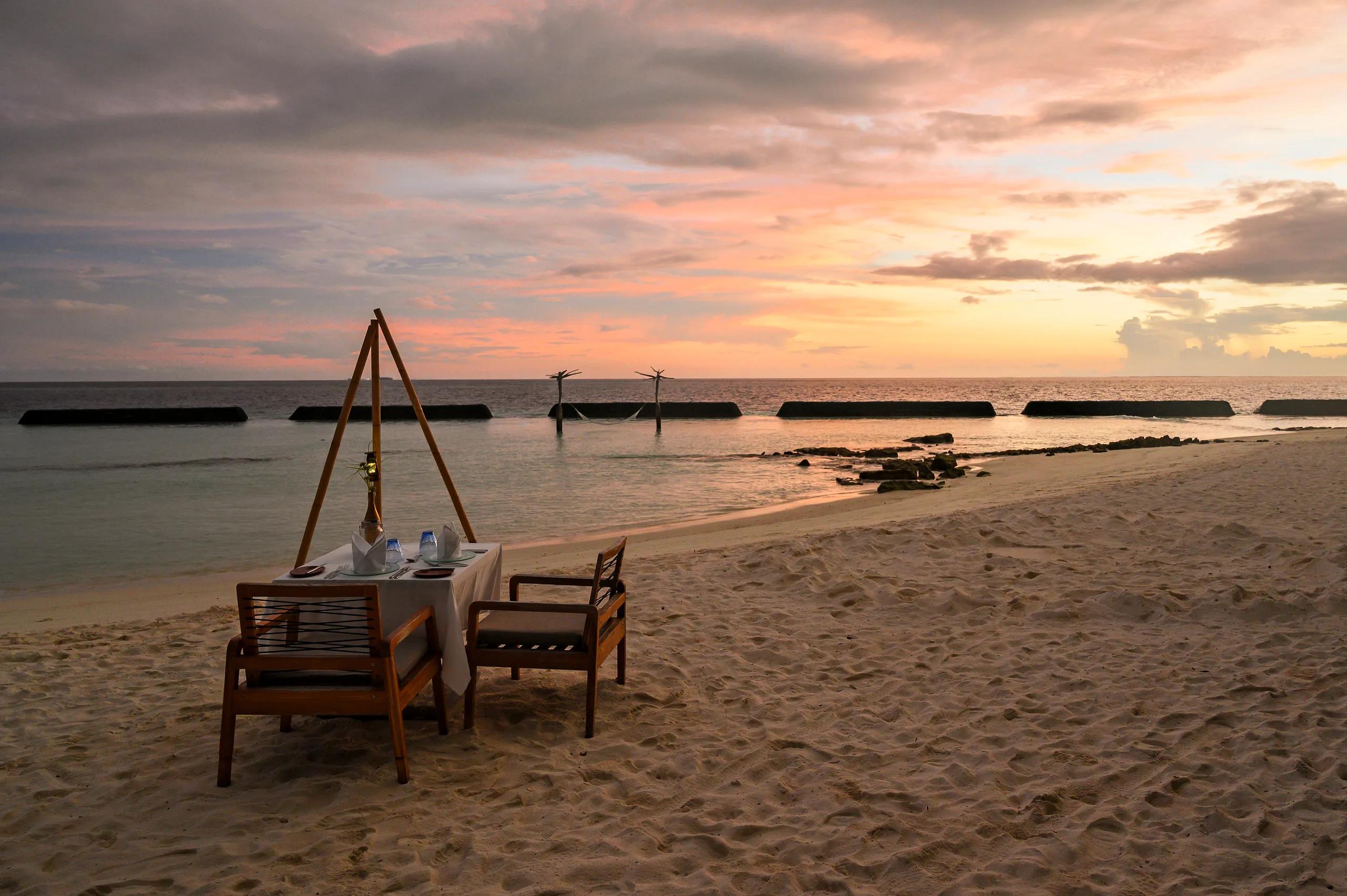 A candlelit table for two set on the sand at the water's edge at dusk, calm lagoon water stretching toward the horizon