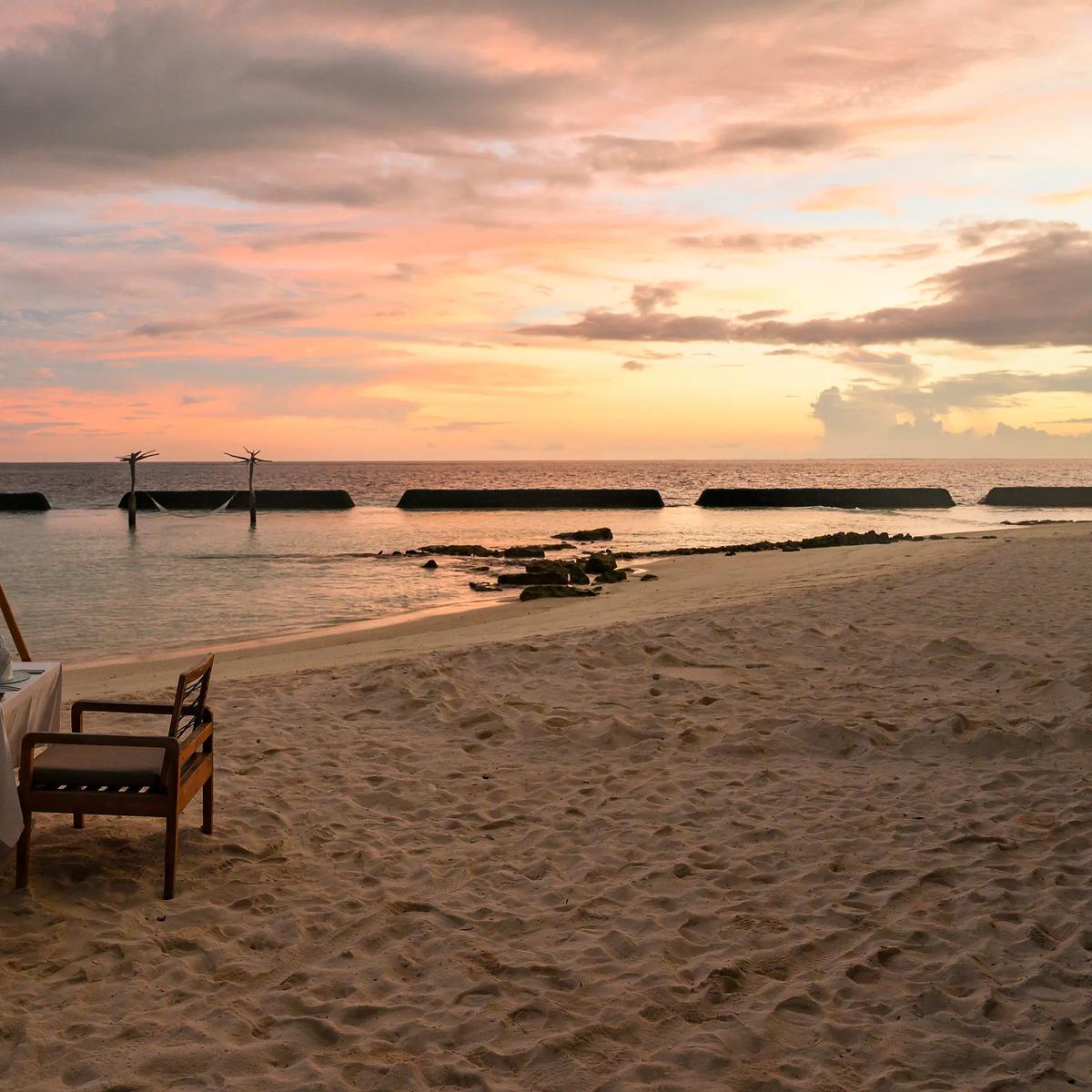 A candlelit table for two set on the sand at the water's edge at dusk, calm lagoon water stretching toward the horizon