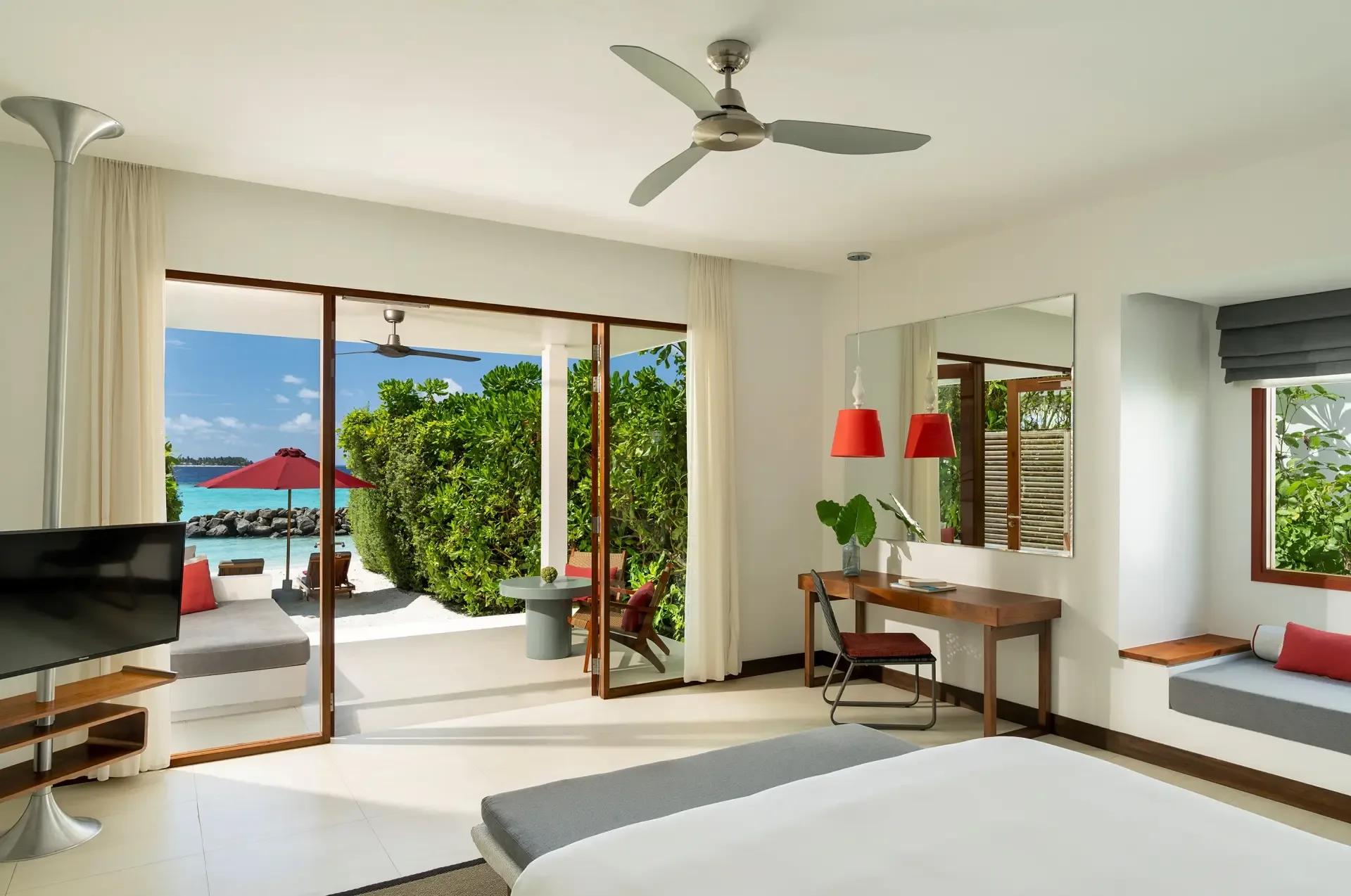 Bedroom of a Beach Villa at Niva Dhigali, sliding doors open to a red-umbrella terrace and the sea beyond