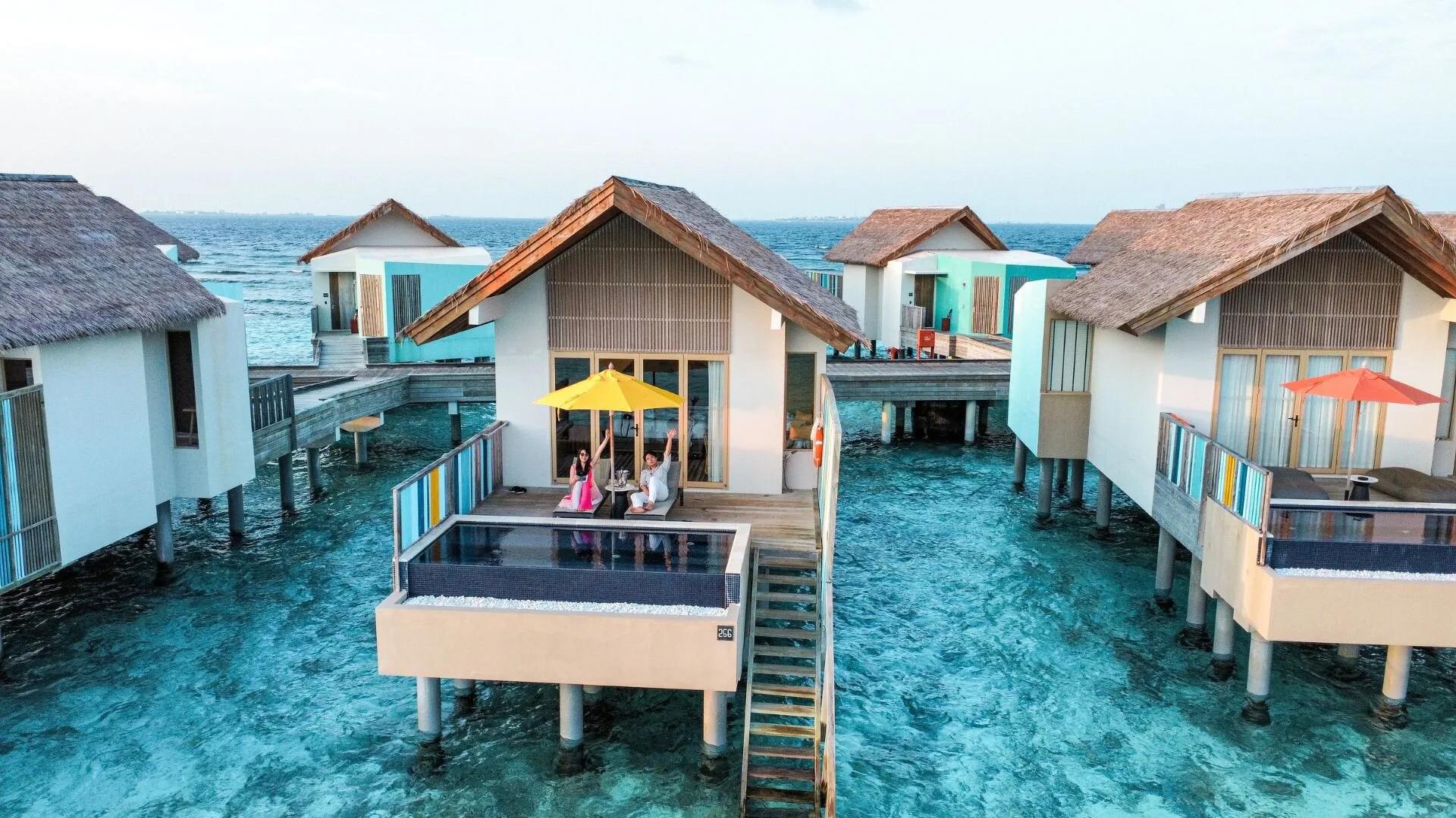 An aerial view of Hard Rock Hotel Maldives on Akasdhoo Island, overwater villas reaching into the lagoon beside the beach