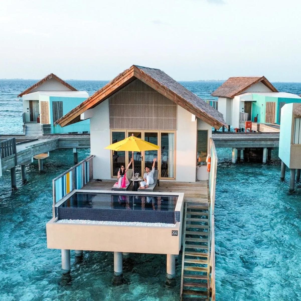 An aerial view of Hard Rock Hotel Maldives on Akasdhoo Island, overwater villas reaching into the lagoon beside the beach
