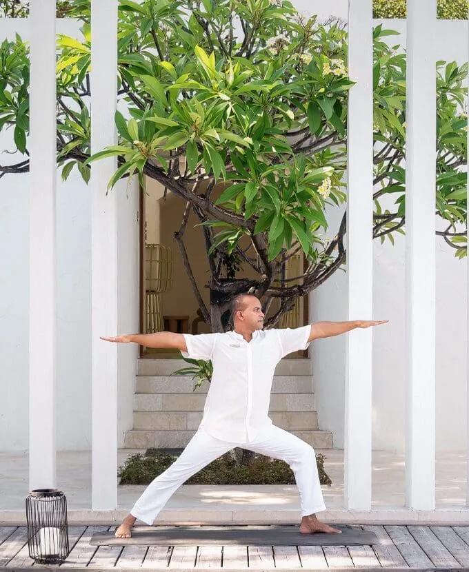 An instructor in white holding a yoga warrior pose on an outdoor deck beneath a frangipani tree