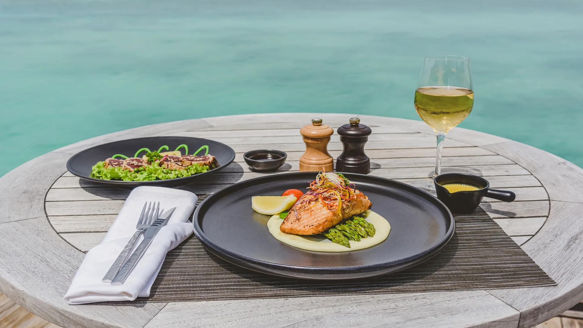 A grilled salmon fillet plated with asparagus and a glass of white wine, the turquoise lagoon in the background at the Grill Bar