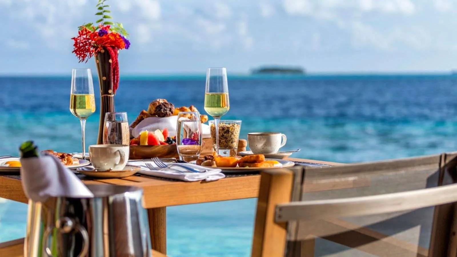 A breakfast spread of pastries, fruit, granola and champagne laid on an overwater villa deck table