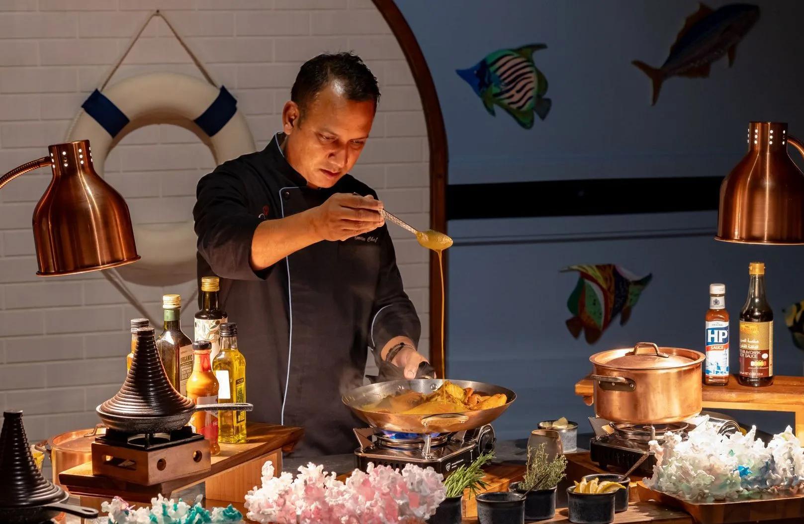 A chef finishing a seafood dish tableside, copper pans and bottled condiments on the counter
