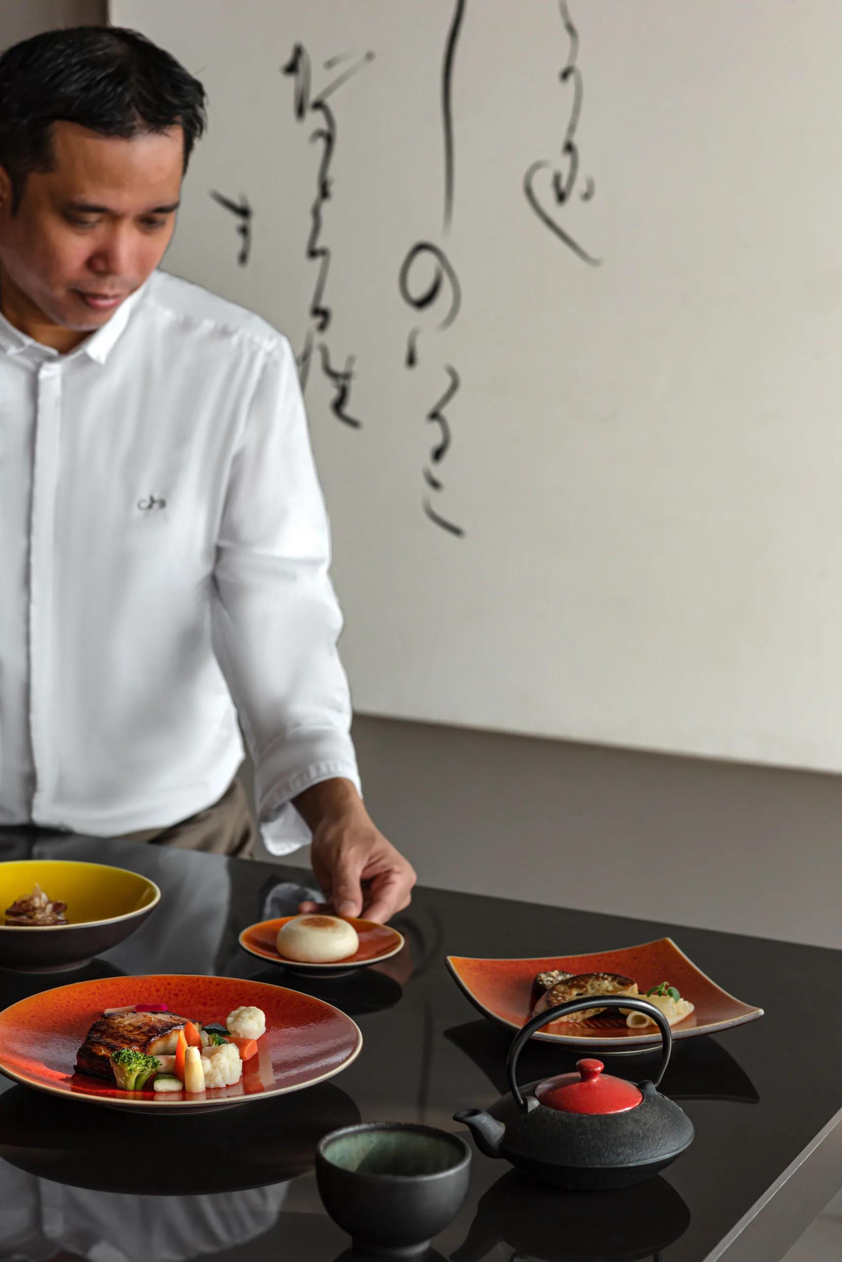A chef at The Diptyque plating a dish beside a calligraphy-painted wall, a diners' table set in the foreground