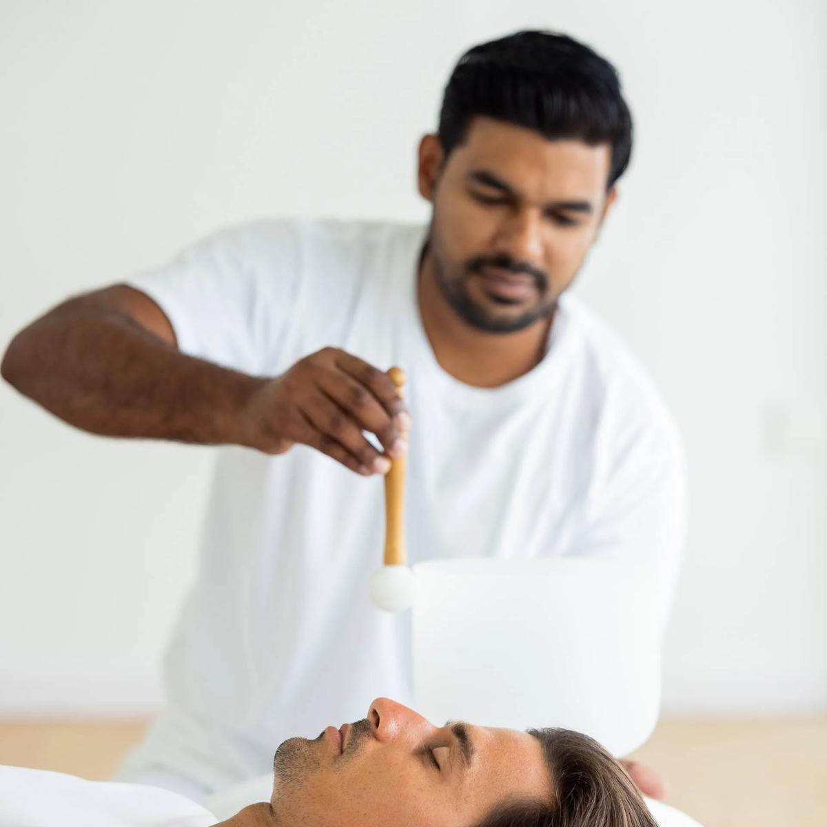 A therapist holding a singing bowl above a reclining guest's face during a sound-healing treatment