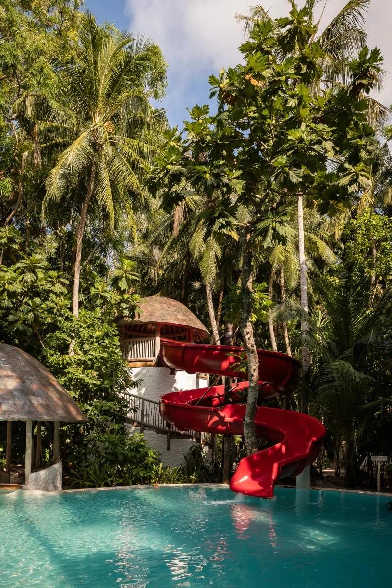 A red spiral waterslide curves down into a pool ringed by mushroom-roofed pavilions and palm trees, part of The Den's grounds