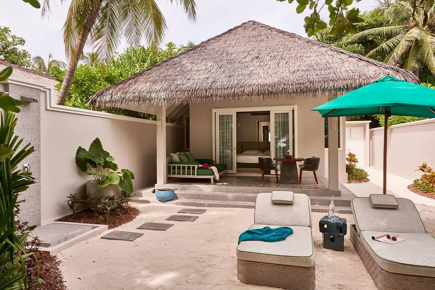 A thatched-roof Beach Villa's private garden terrace, with a green daybed sofa and loungers under a blue umbrella