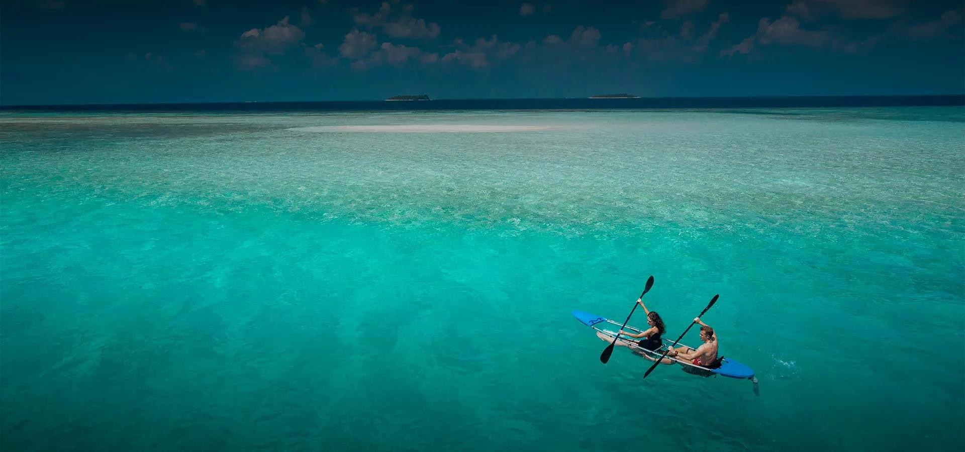 A couple paddling a clear-hulled kayak across Baros's shallow lagoon, a sandbank and the reef edge visible in the turquoise water beneath them