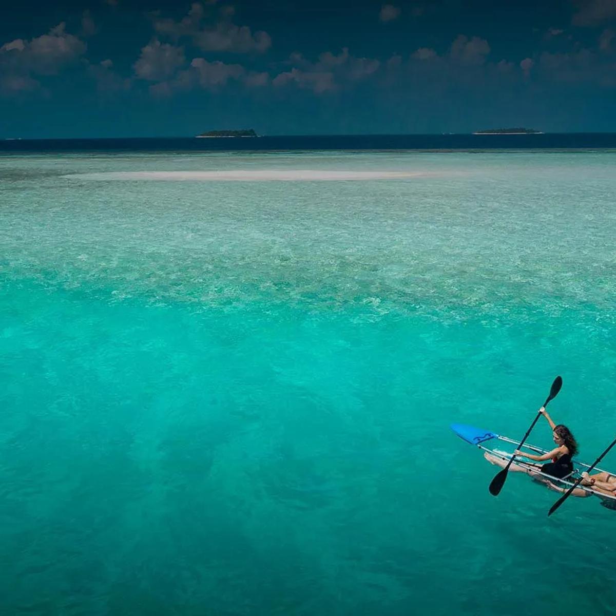 A couple paddling a clear-hulled kayak across Baros's shallow lagoon, a sandbank and the reef edge visible in the turquoise water beneath them