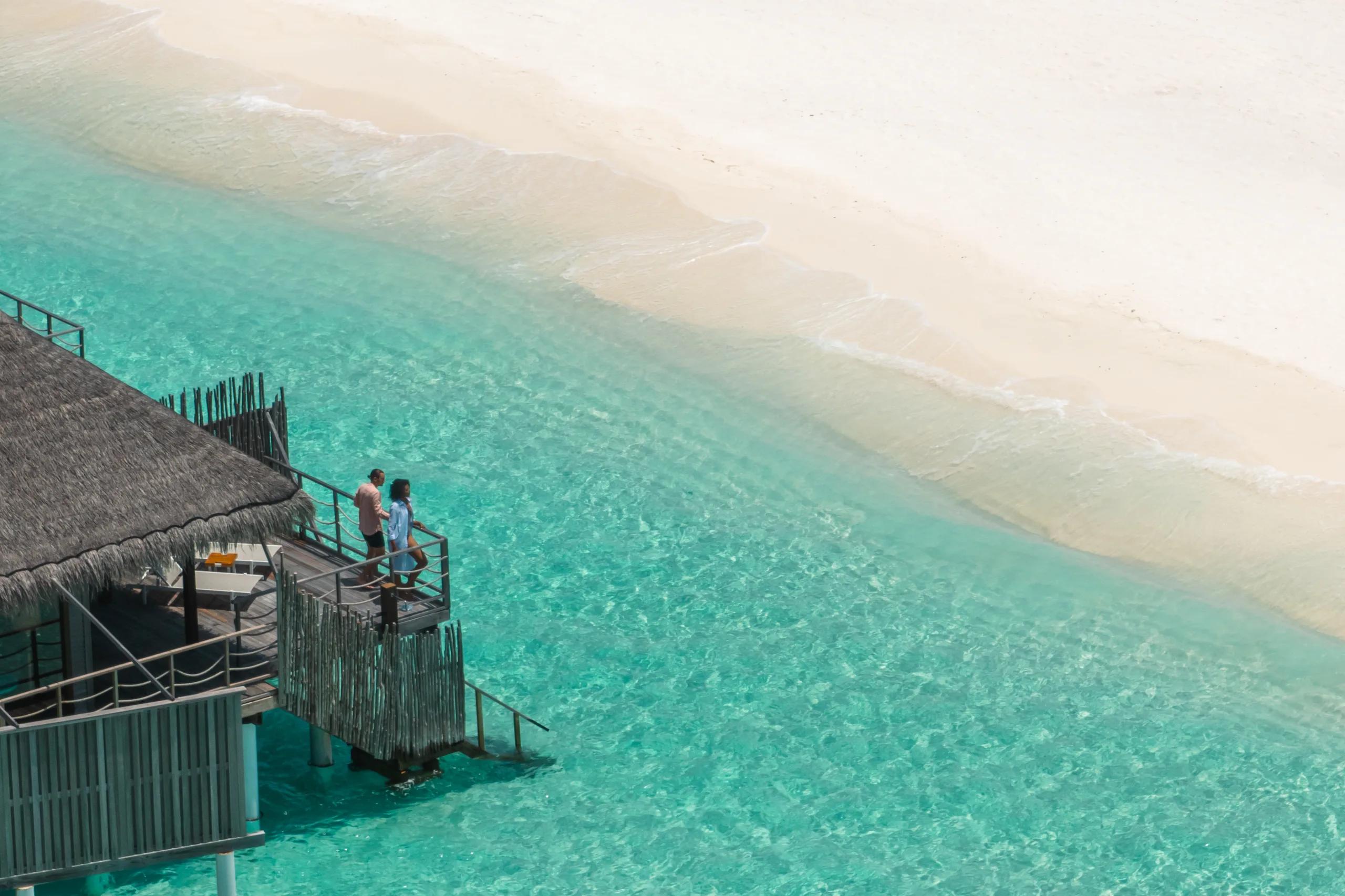 Aerial view of a couple on the steps of a water villa jetty at dusk, white beach on one side and open lagoon on the other