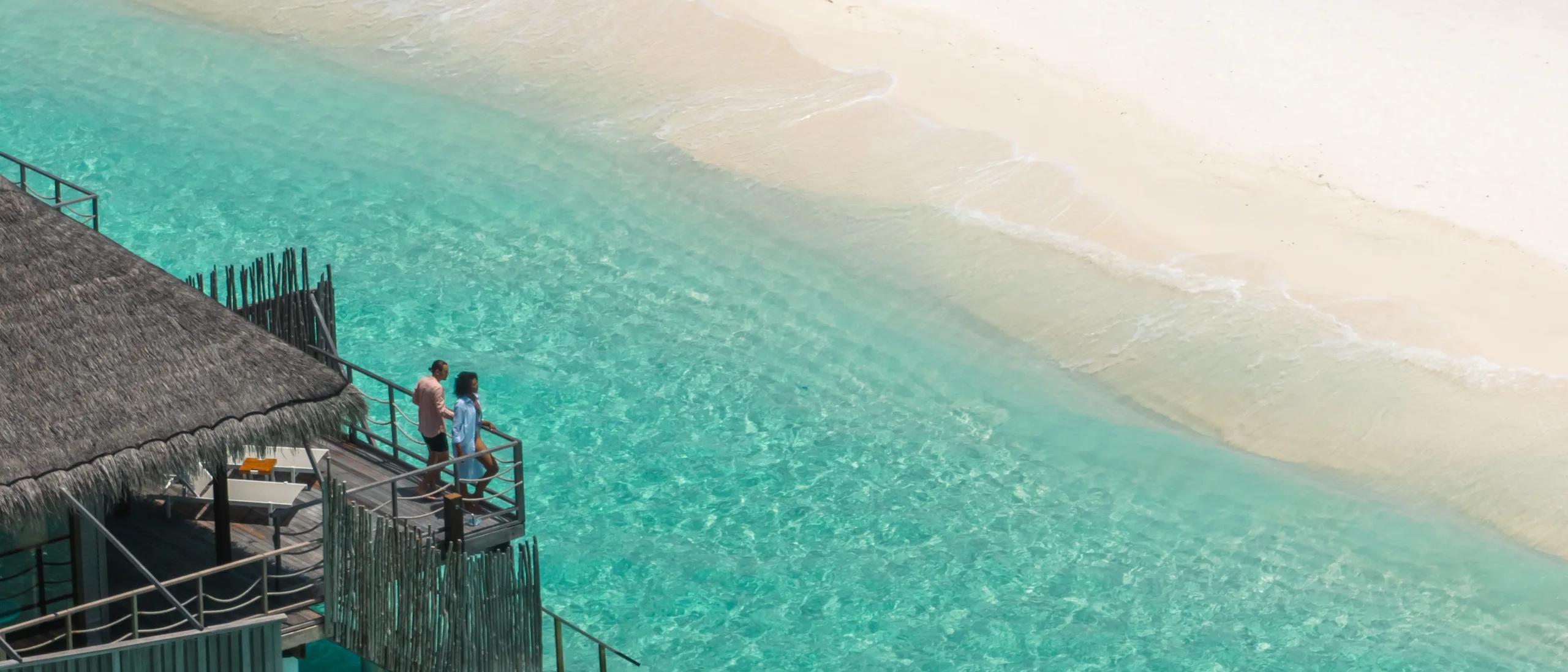 Aerial view of a couple on the steps of a water villa jetty at dusk, white beach on one side and open lagoon on the other