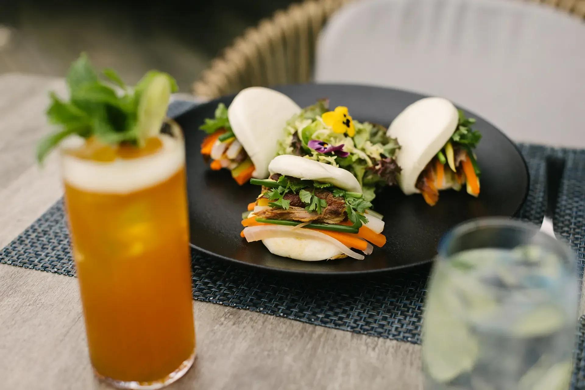 Steamed bao buns filled with meat and vegetables, plated beside a garnished iced cocktail