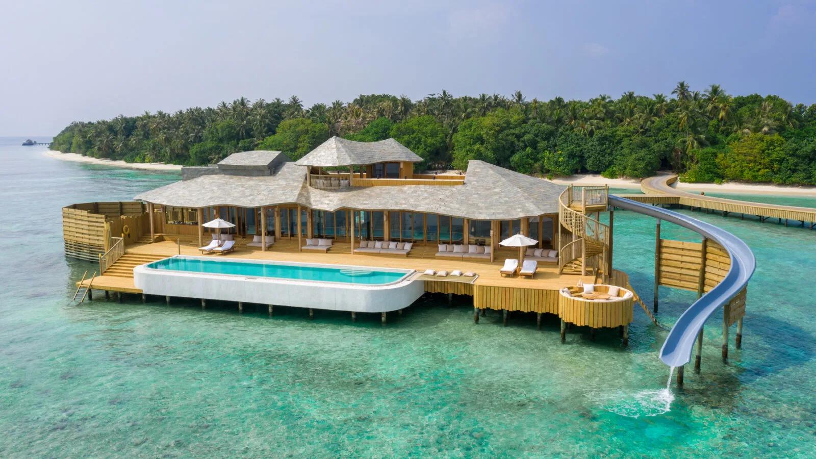 A 2-Bedroom Water Reserve with Slide, its white pool built out over the reef with a slide curving down into the lagoon and the island's beach visible behind