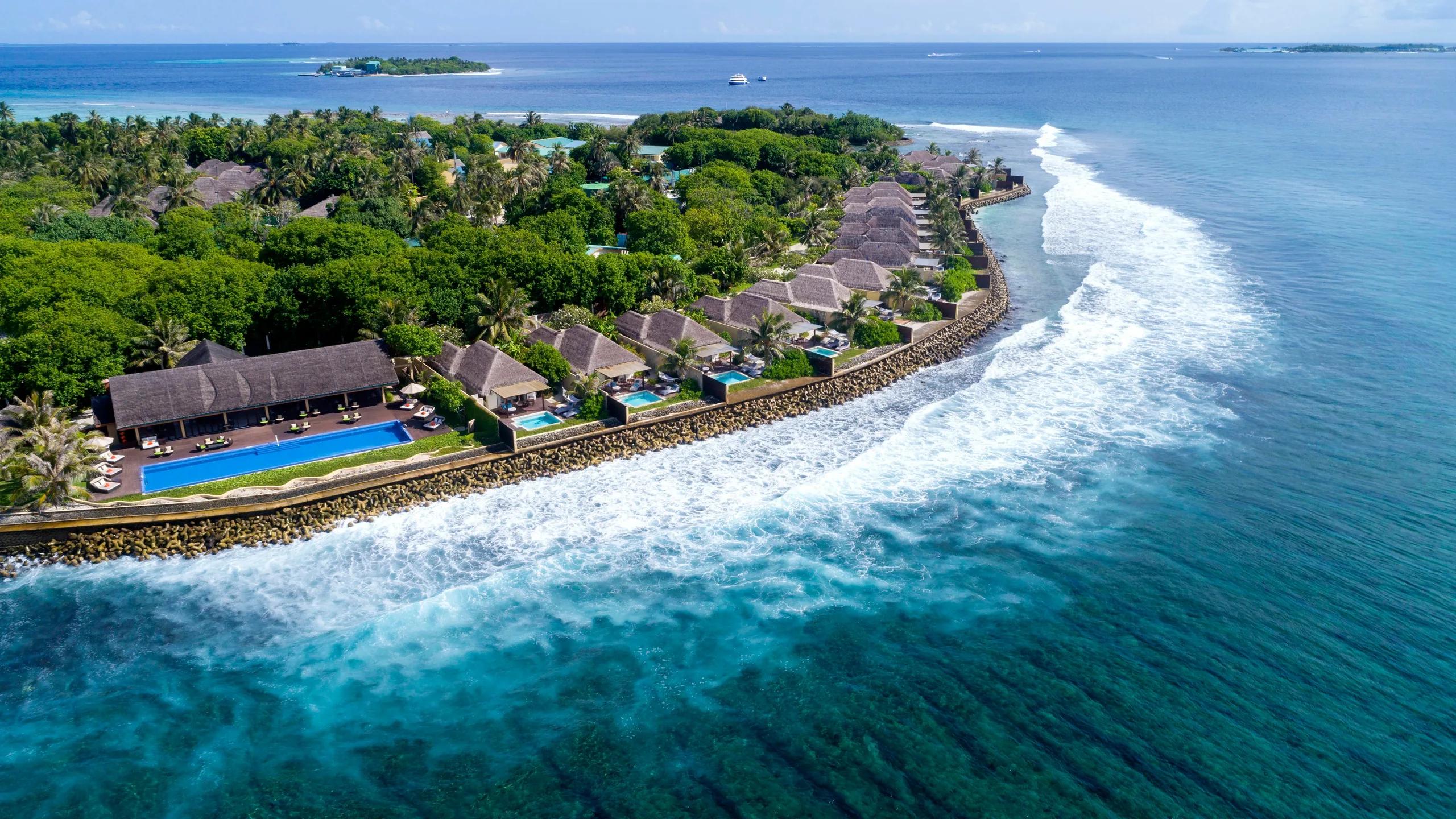 Aerial view of the island's ocean-facing coastline, with a row of villas and a swimming pool set behind the reef break