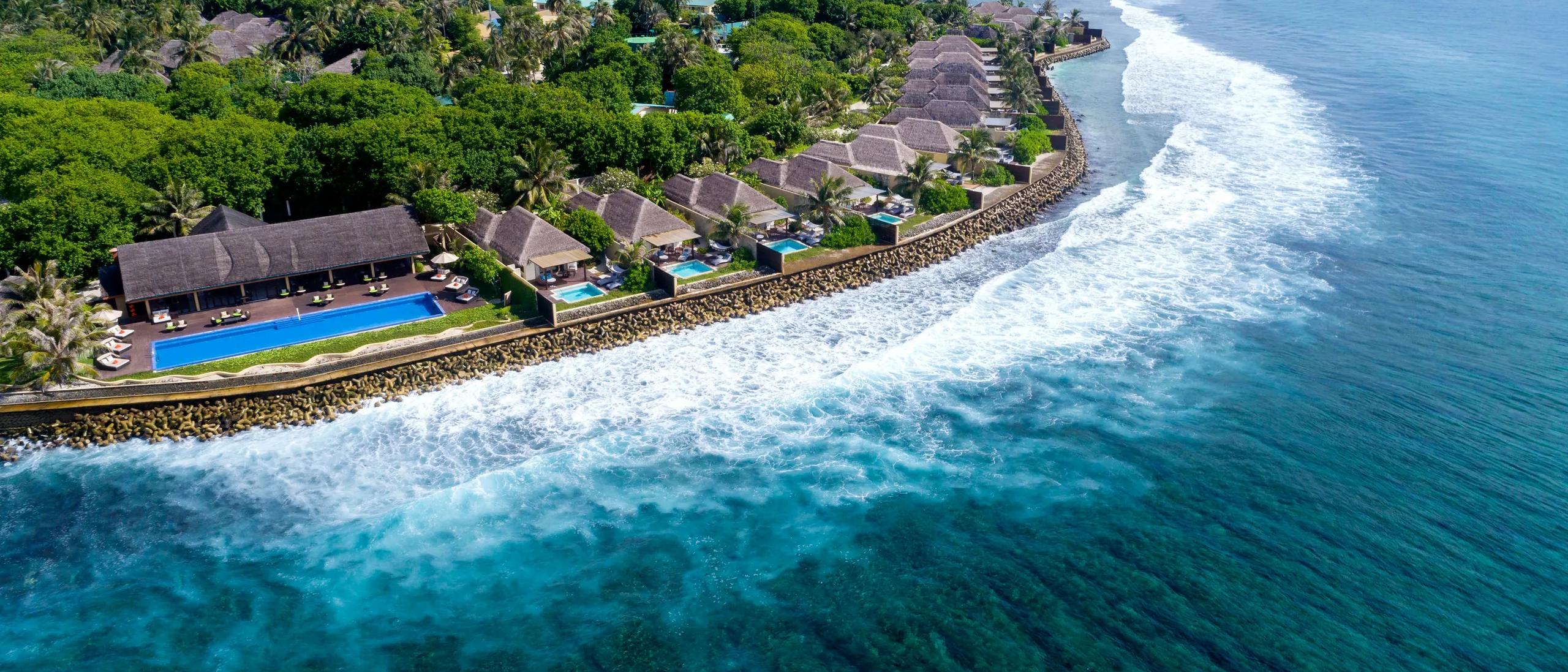 Aerial view of the island's ocean-facing coastline, with a row of villas and a swimming pool set behind the reef break